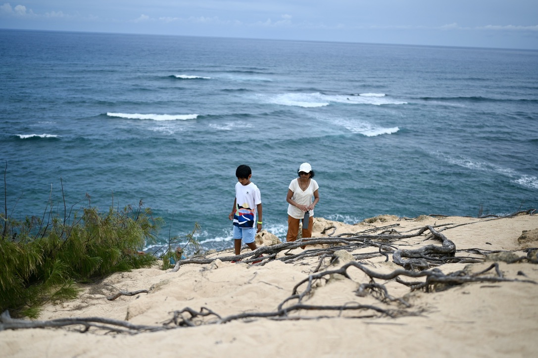 _Boy and elder woman on the cliff side of a sandy beach with roots and ocean with light waves behind them