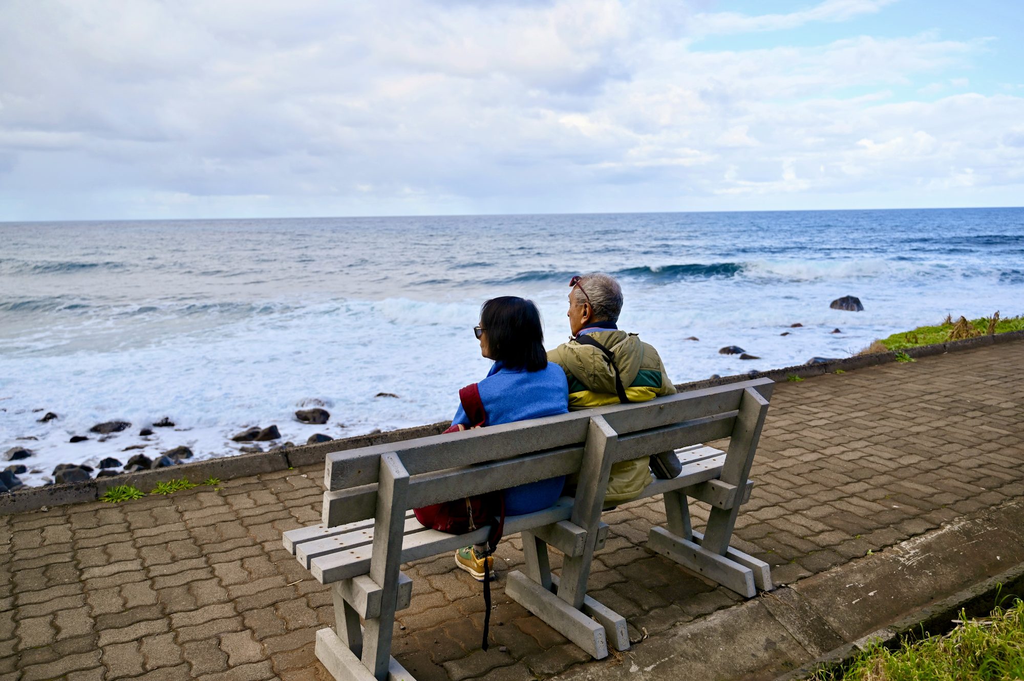 Two of us sitting on a wooden bench on the seafront path, looking out at the waves breaking over black rocks.