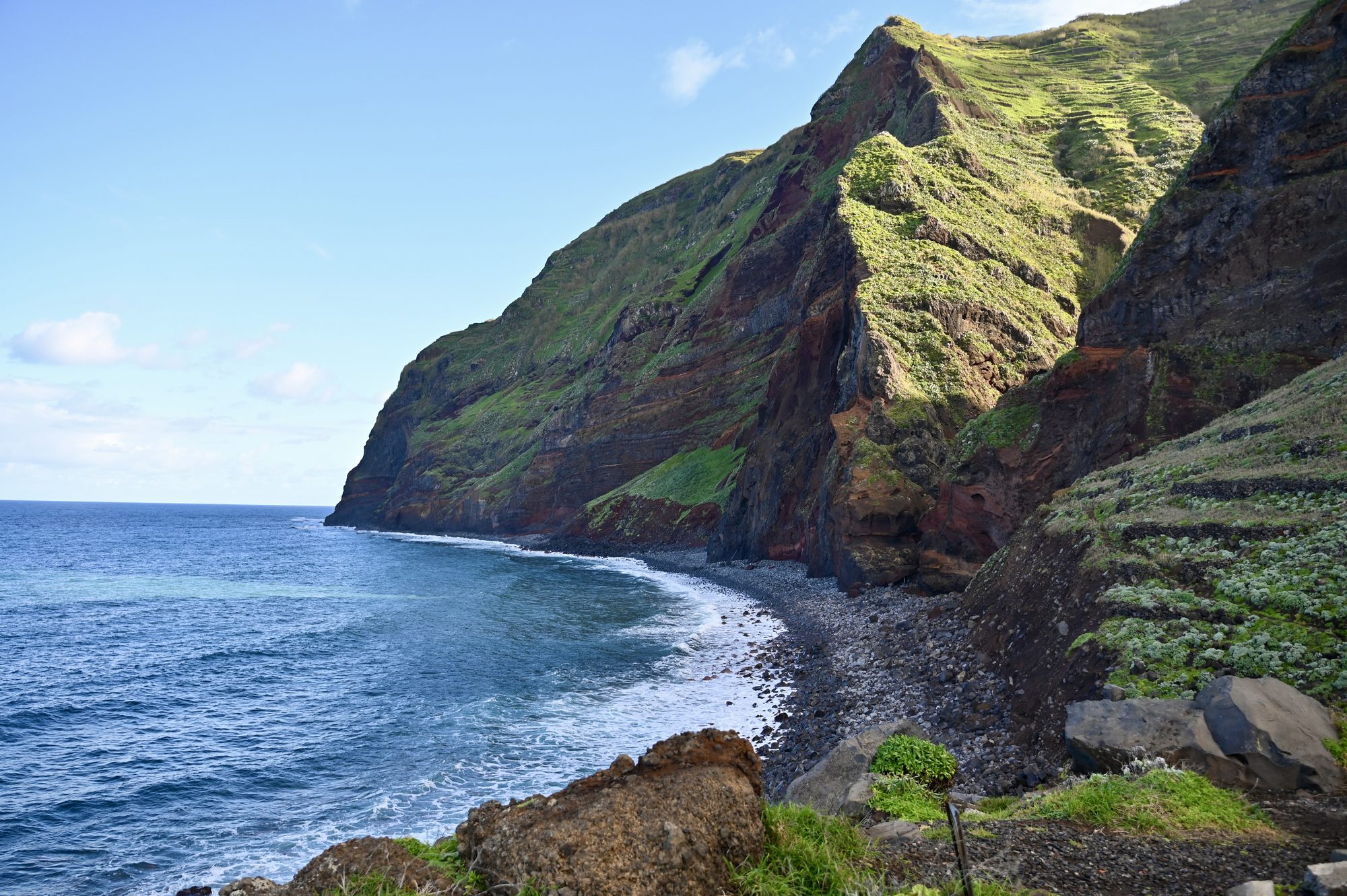 A towering green cliff dropping to a dark pebble beach and blue sea.