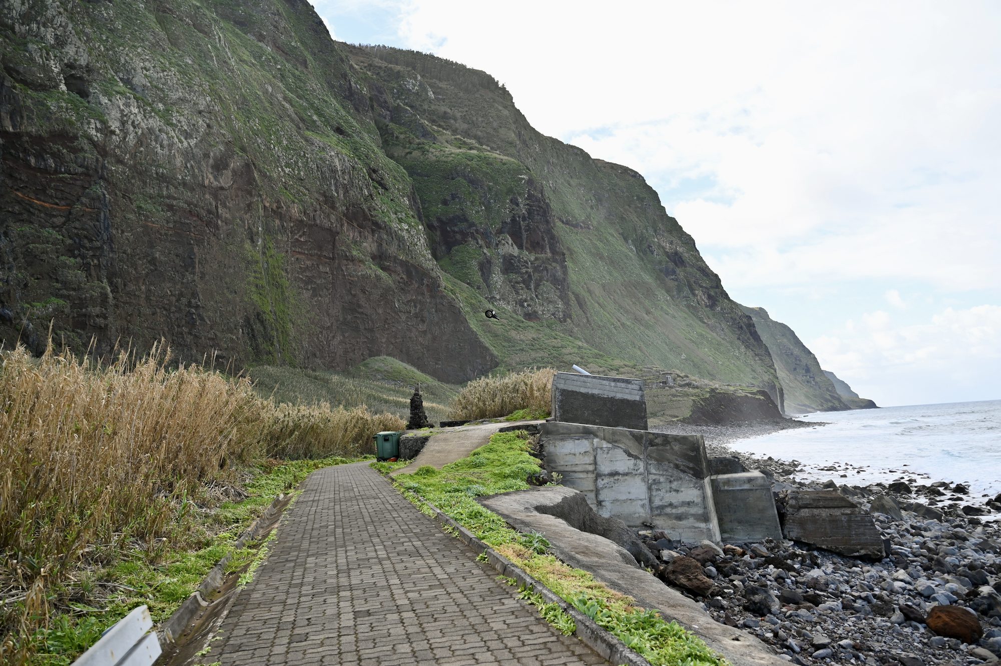 A paved path running along the shore below enormous cliffs, past tall dry grasses and a few small buildings.