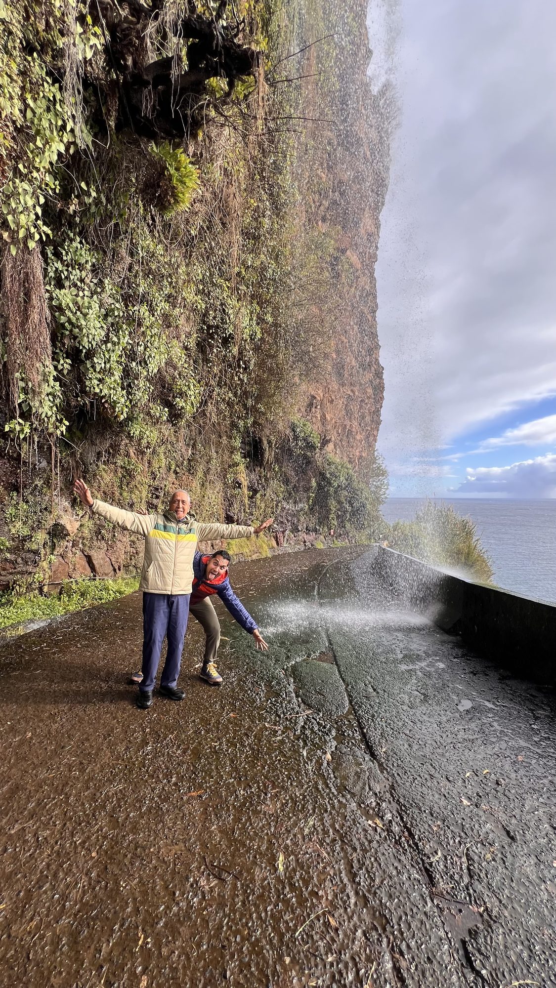 Two of us with arms outstretched on a wet road, where water pours off the cliff above and sprays over the edge towards the sea.