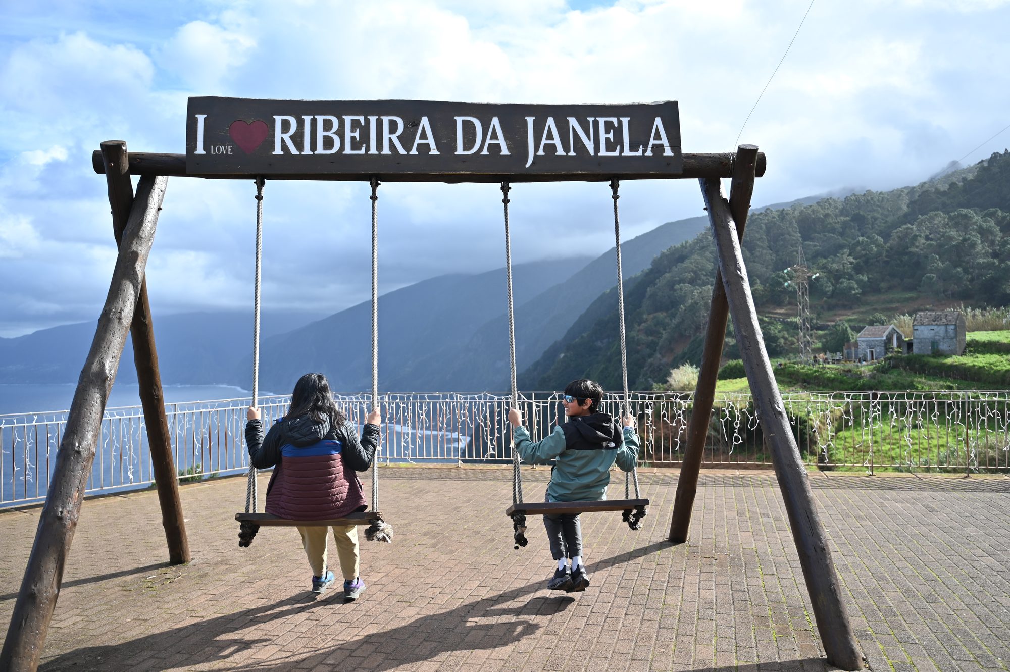 Two of us on wooden swings under an “I love Ribeira da Janela” sign, facing the mountains and the sea.