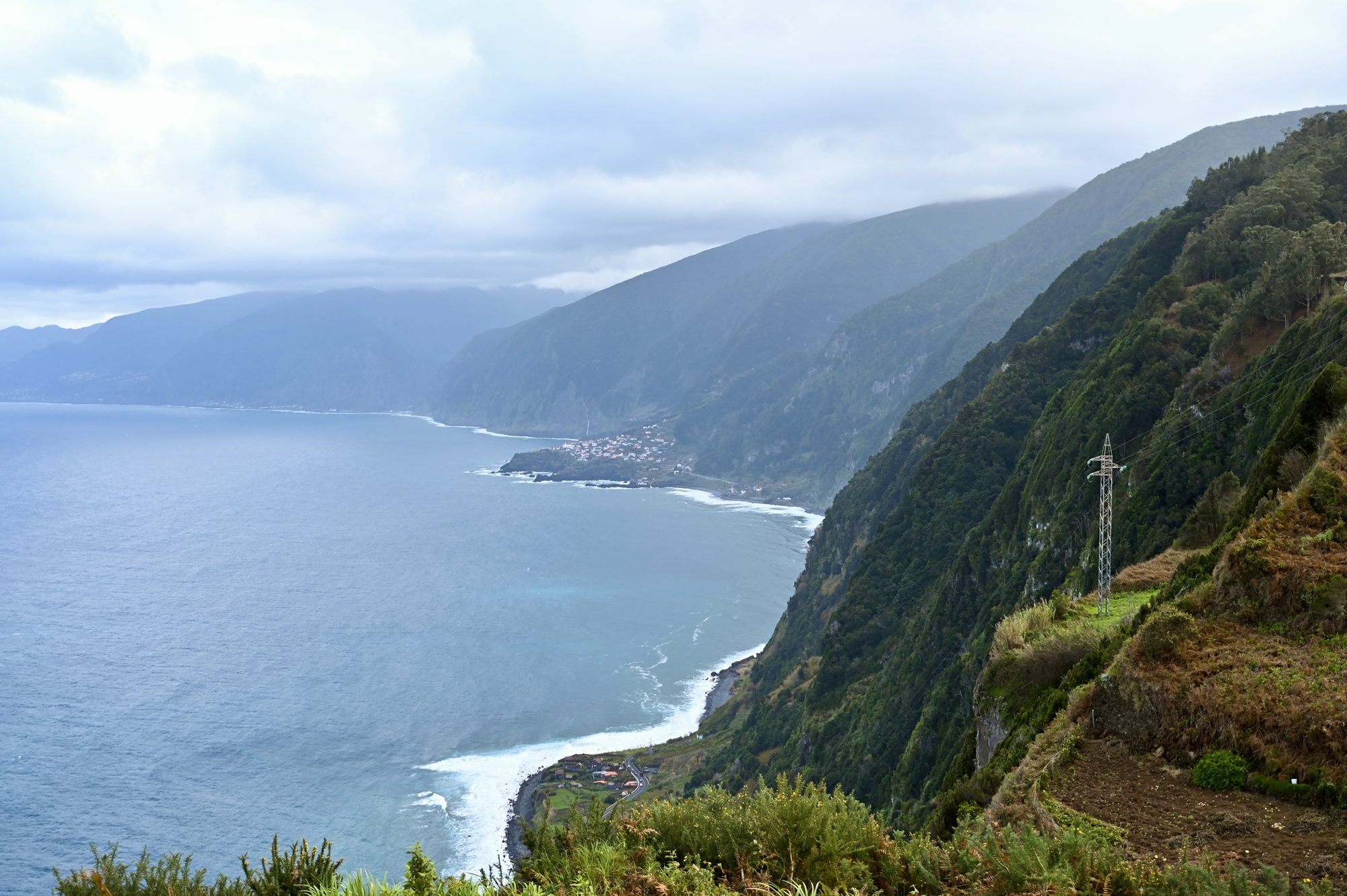 Steep, forested green cliffs falling to a long coastline, with a small town at the water’s edge in the distance.