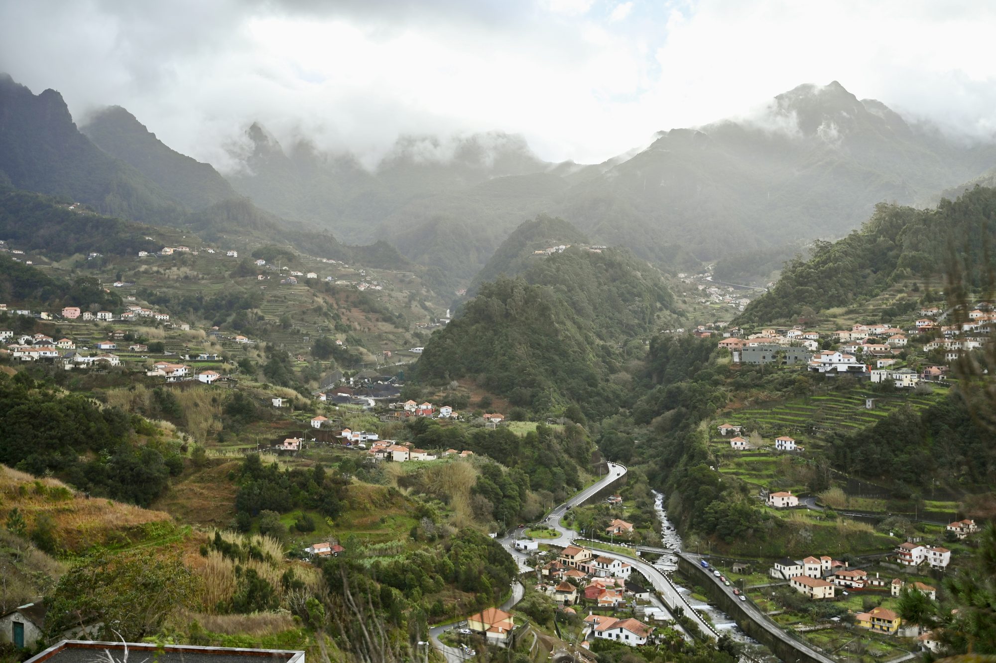 A misty green valley dotted with houses and terraces, with a road winding along the valley floor below the mountains.
