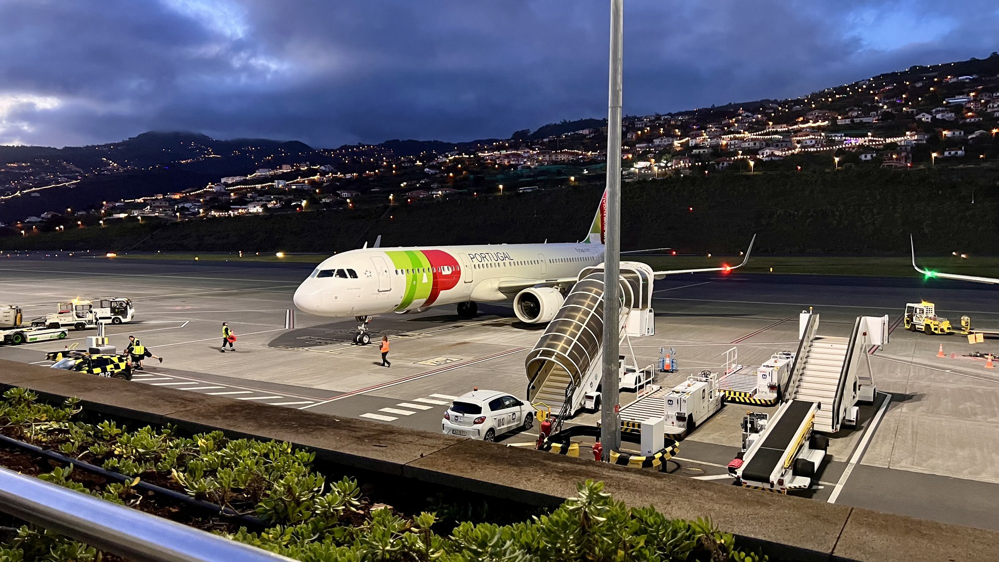 A TAP Air Portugal plane at the gate at dusk, with the boarding stairs in place and the town lit up on the hillside behind.
