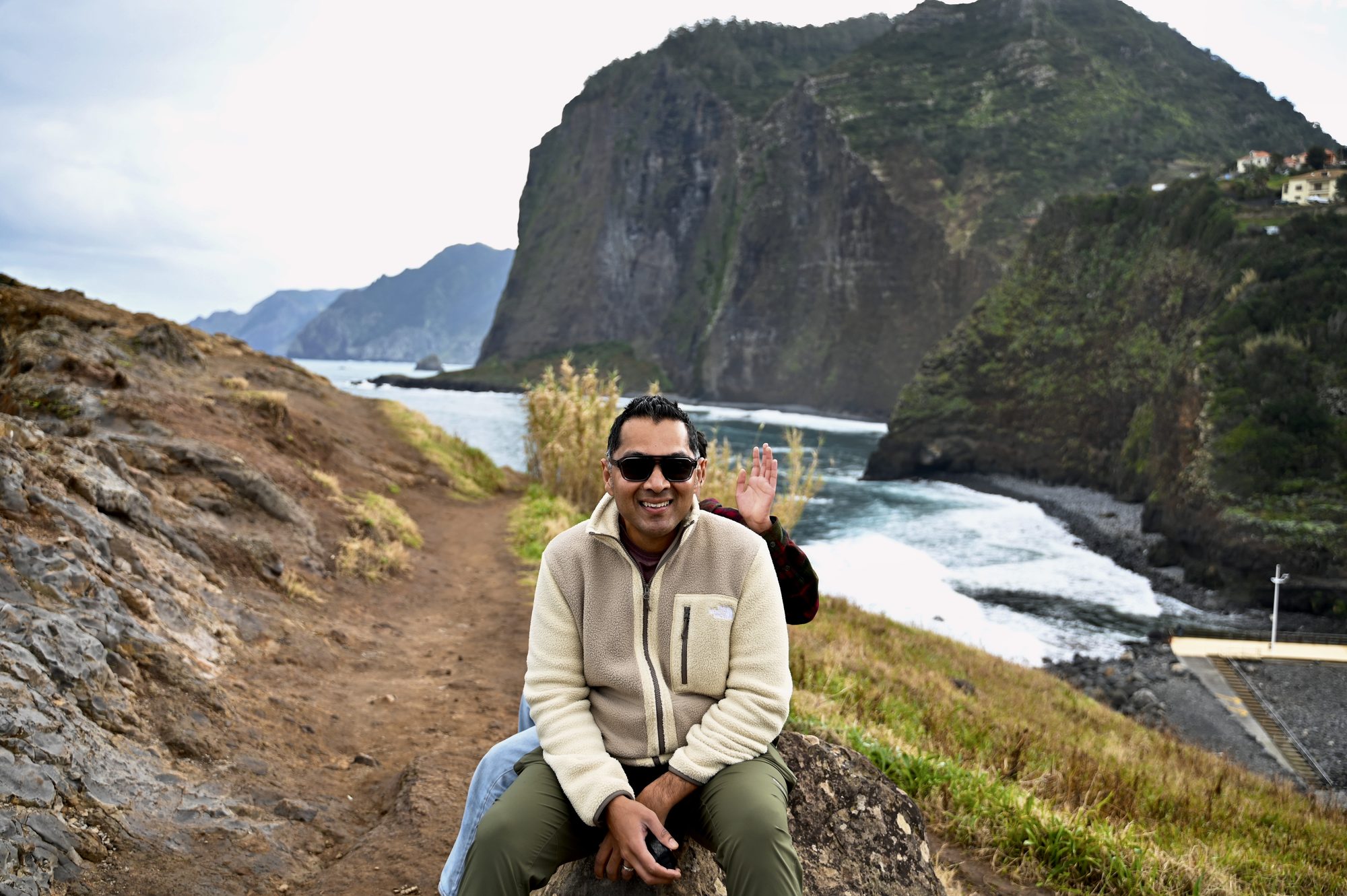 His dad sitting on a rock beside a dirt path, with a huge green headland and the sea behind him.