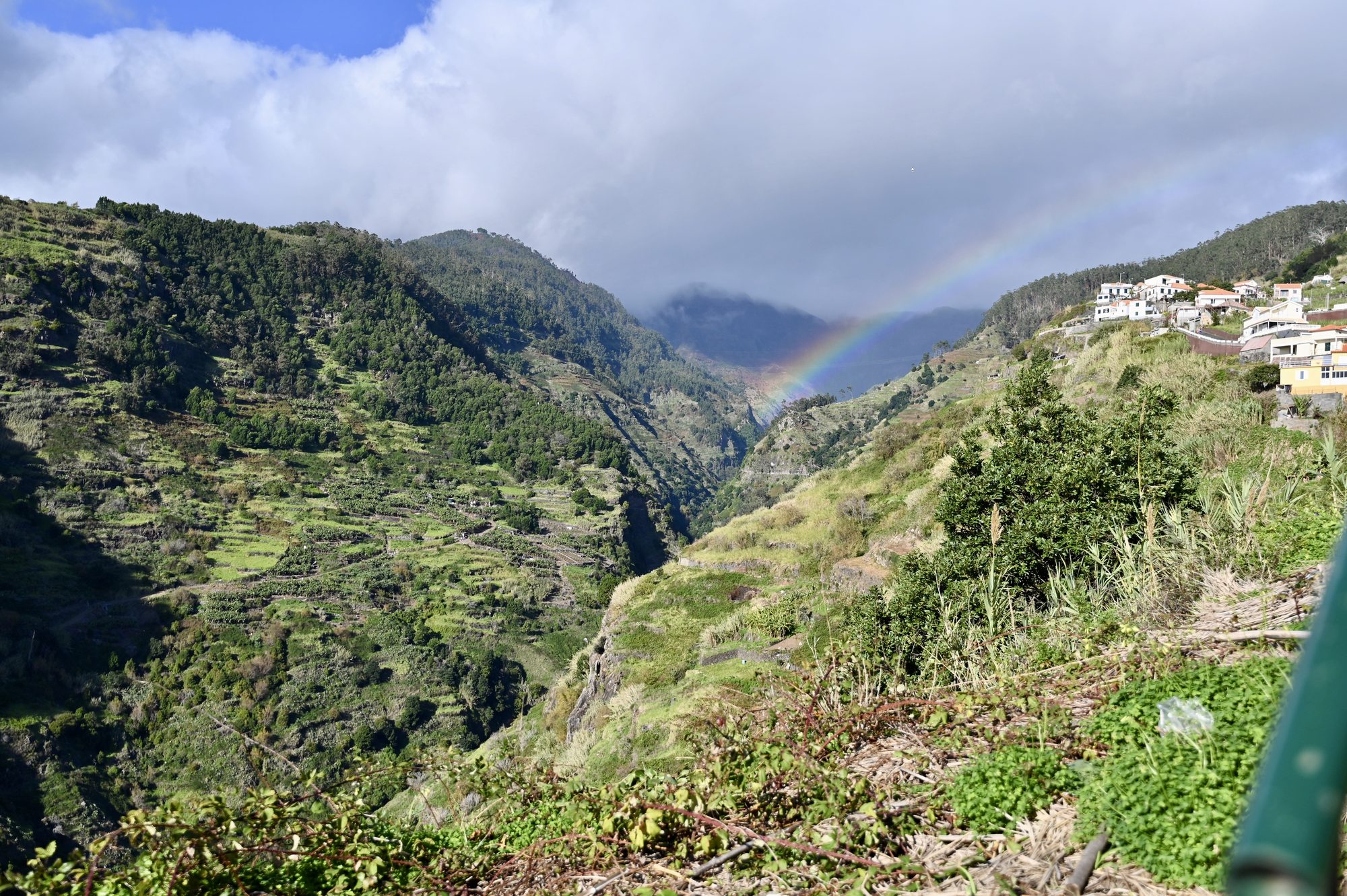 A rainbow arching over a steep, terraced green valley, with white houses on the ridge.