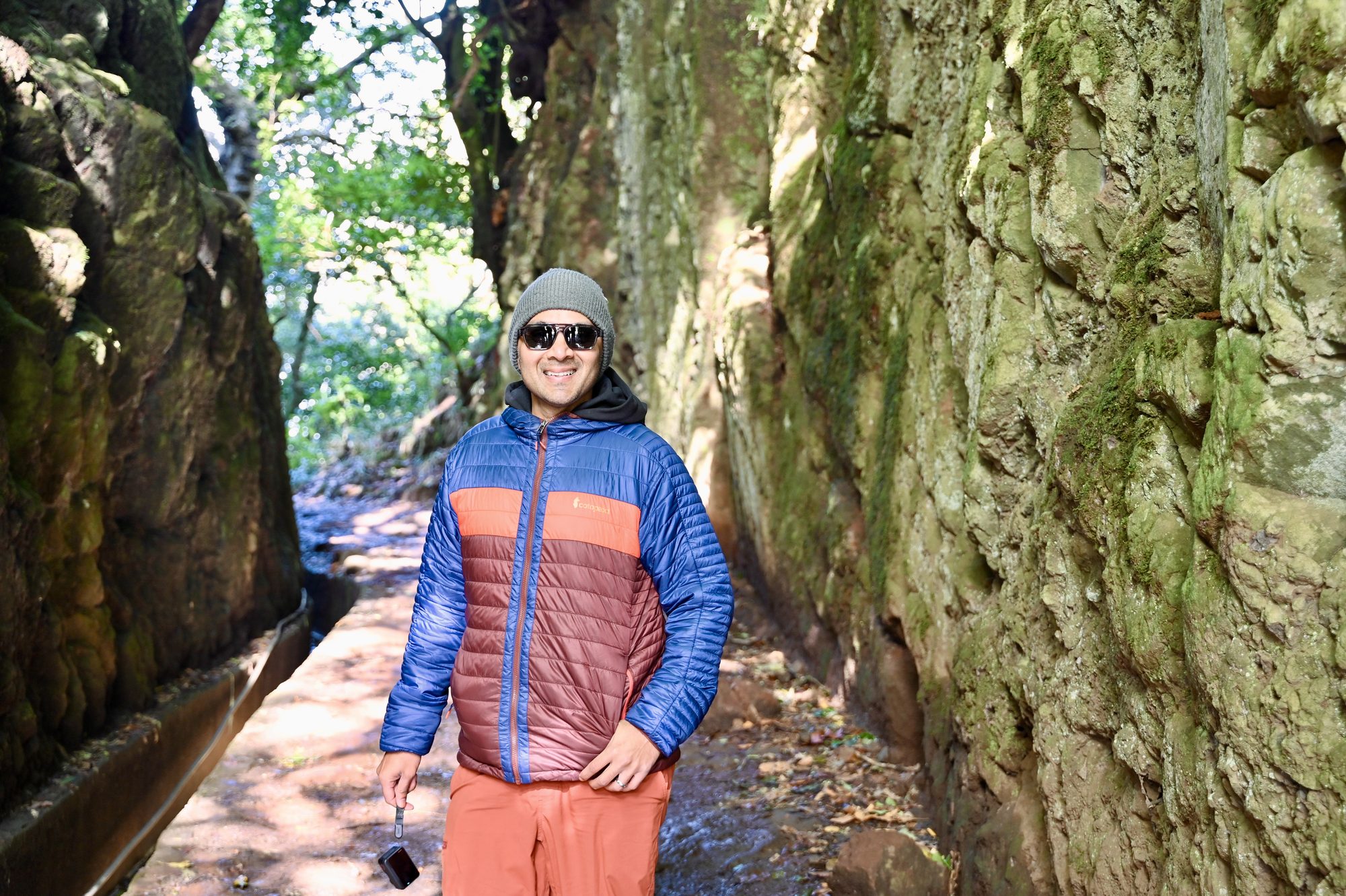 His dad smiling in a padded jacket and beanie on the path between two tall, mossy rock walls beside the levada channel.