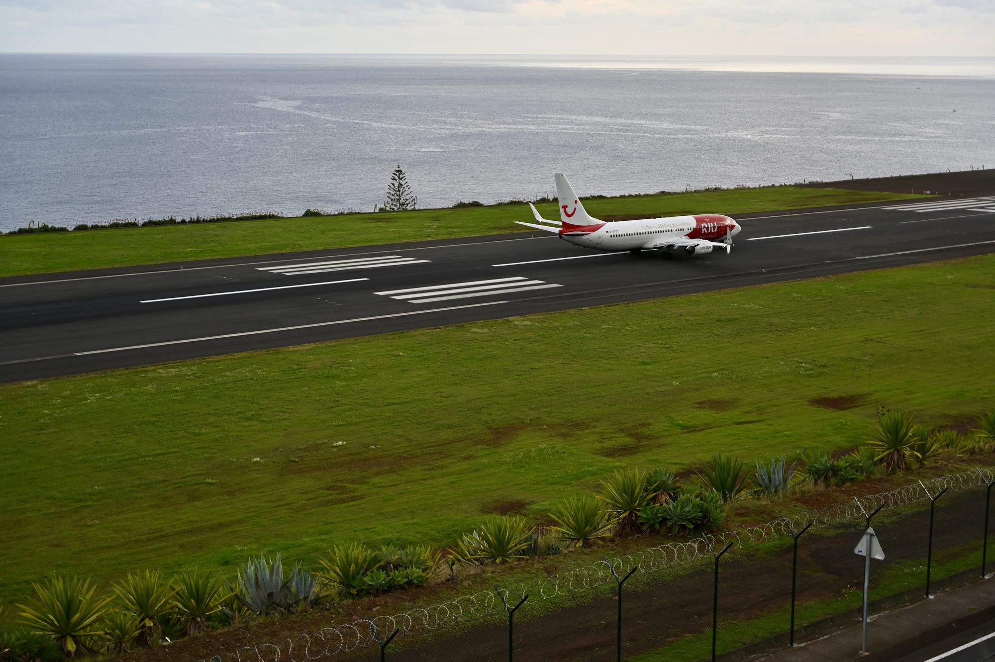 A red and white plane on the runway, with the ocean beyond and a chain-link fence in the foreground.
