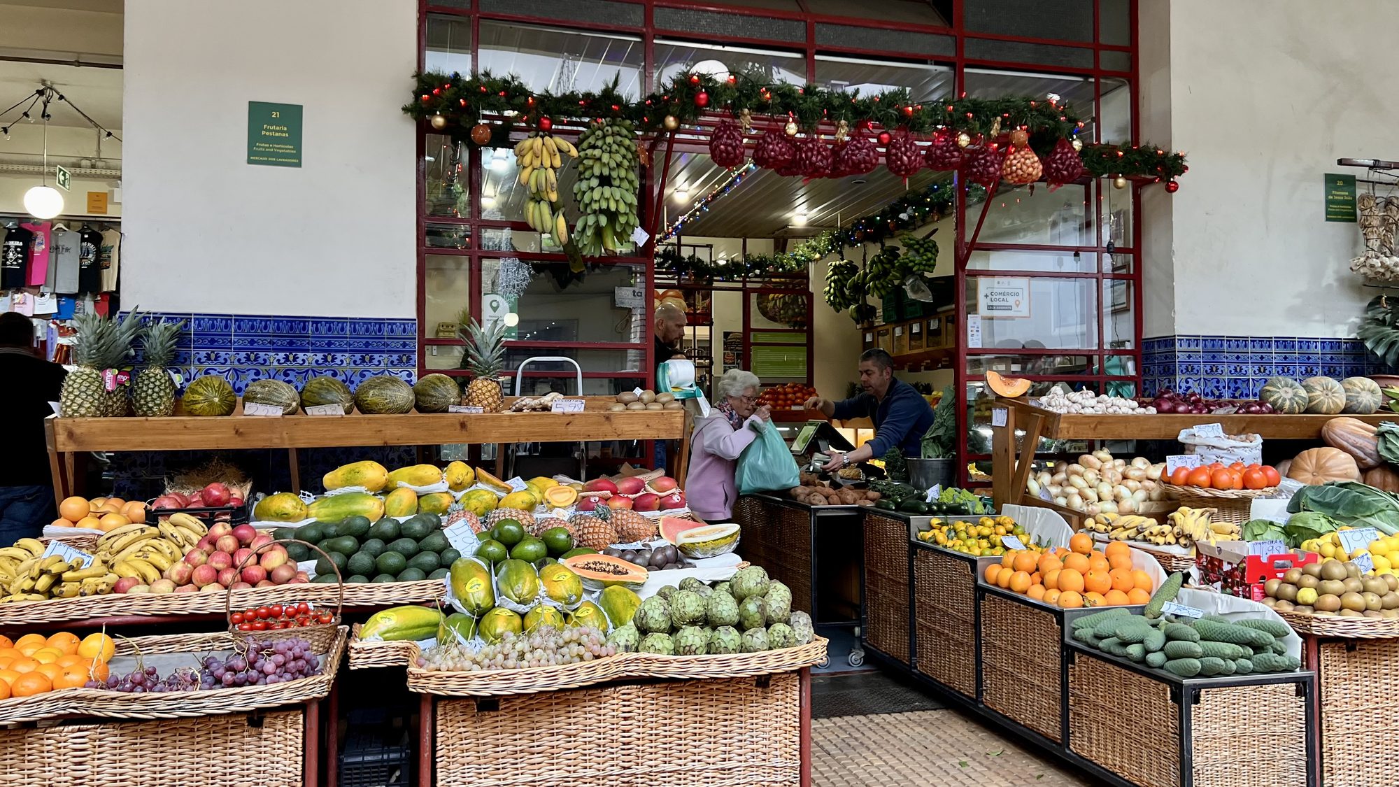 A market stall piled high with tropical fruit in wicker baskets, under Christmas garlands.