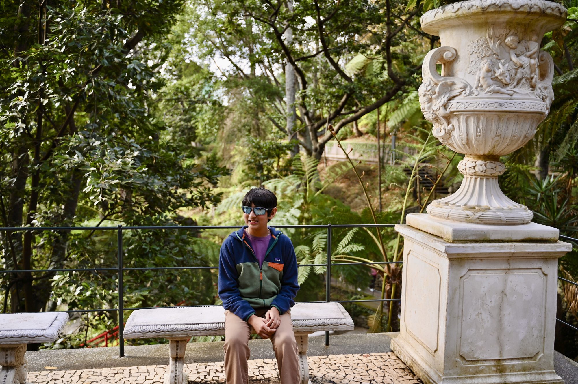 Our son sitting on a stone bench next to a large carved stone urn, among tropical trees.