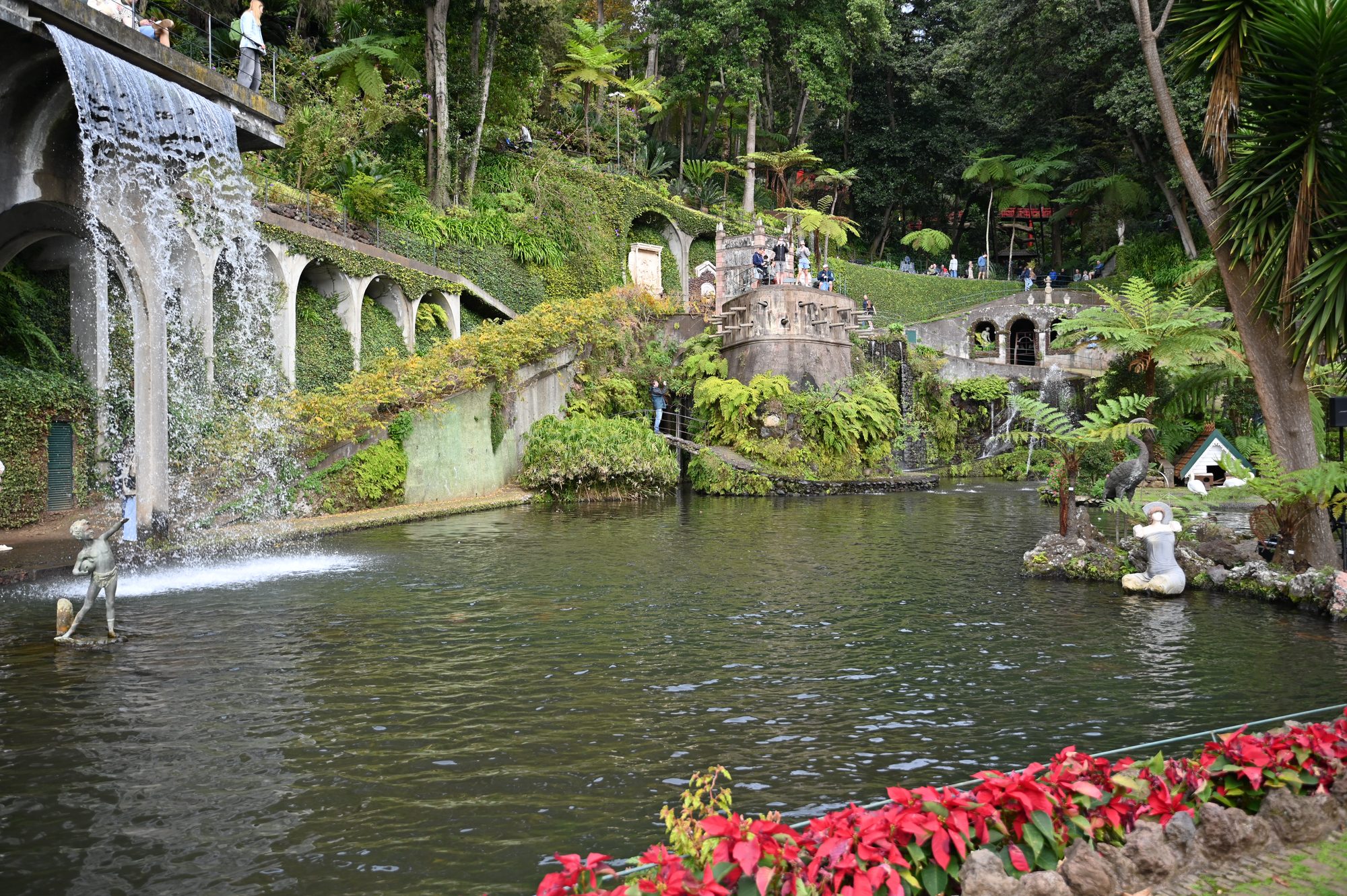 A waterfall spilling from an arched stone wall into a large pond, with red poinsettias along the edge.