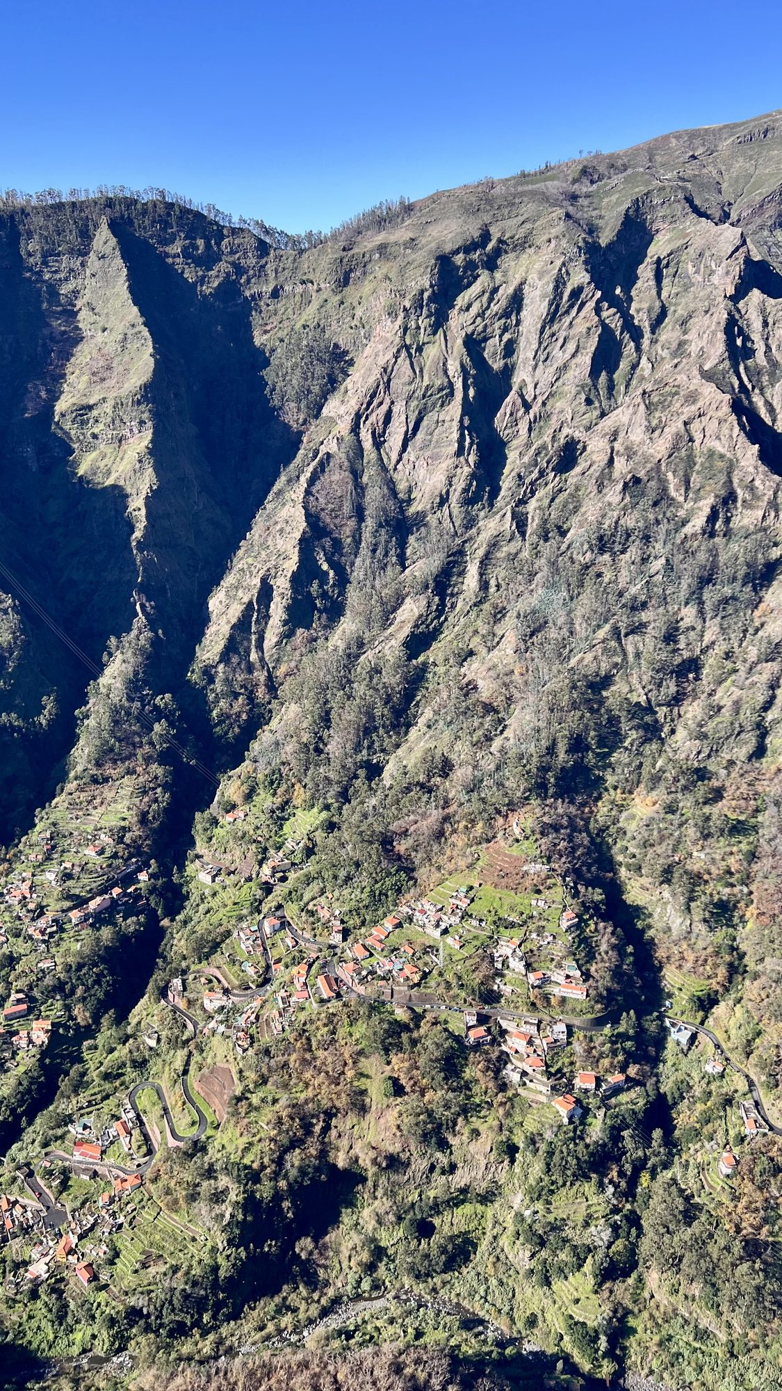 Steep mountain slopes plunging into a valley, with a village of small houses and terraces on the valley floor.