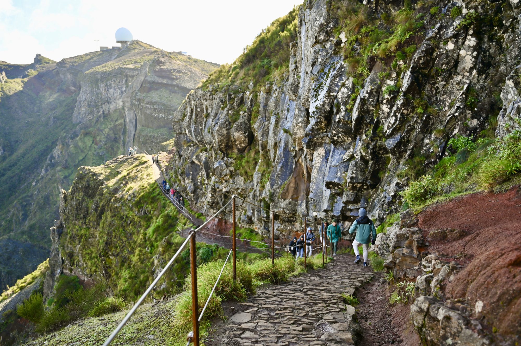 Hikers on a cobbled path along the cliffside, with a cable railing and the white observatory dome on the summit behind.