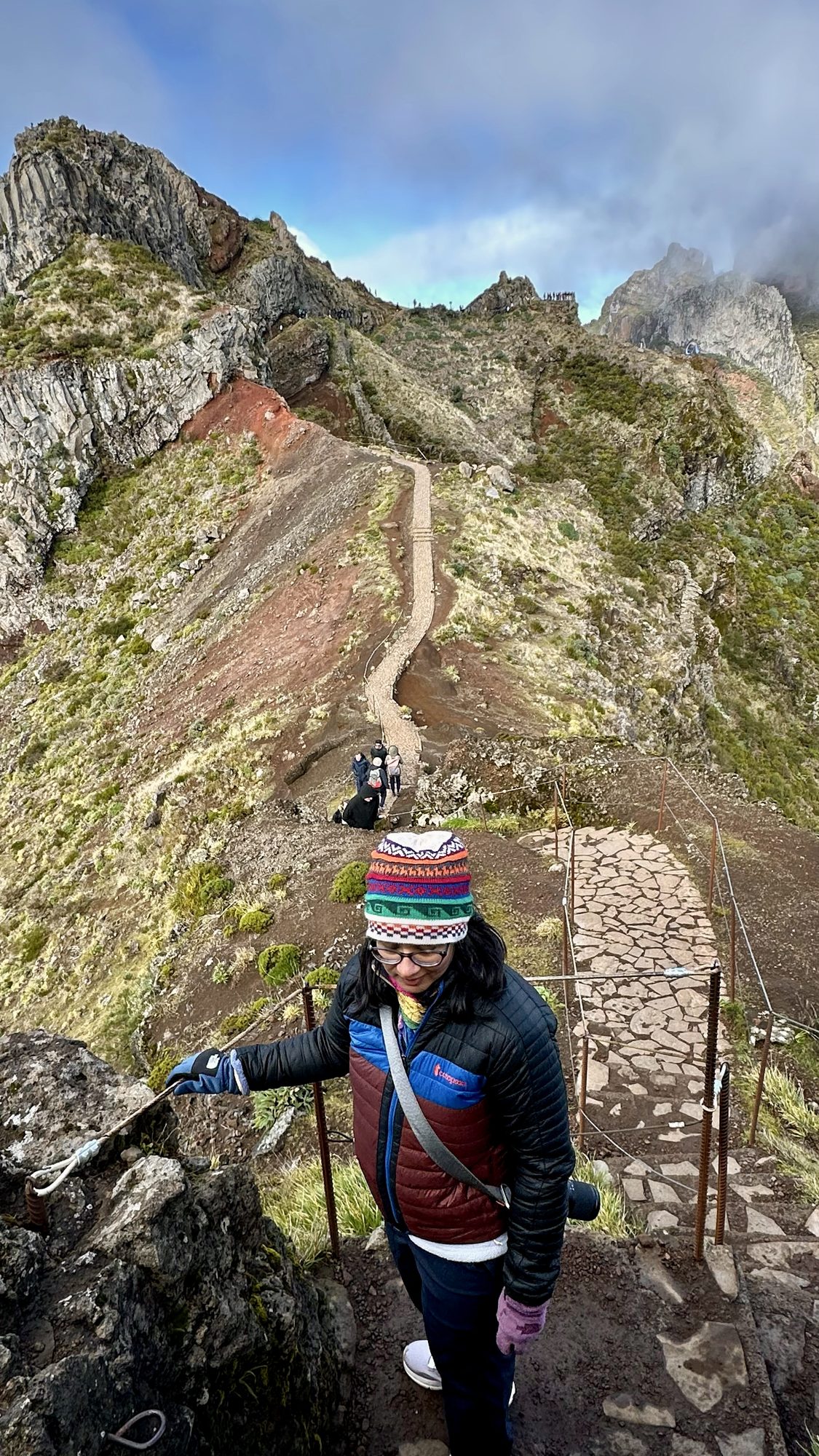 One of us in a woolly hat holding the cable railing on a narrow stone path that winds along a red-earth ridge.