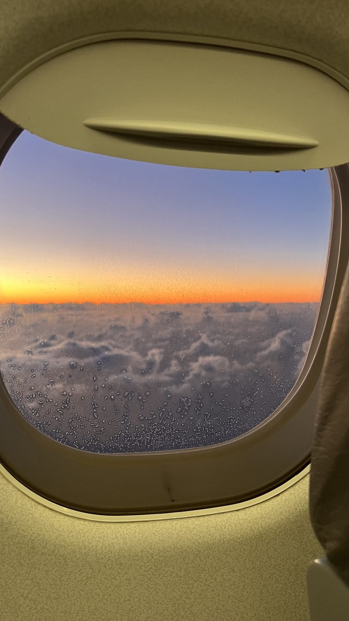 The sky glowing orange at sunrise above a layer of clouds, seen through an aeroplane window.