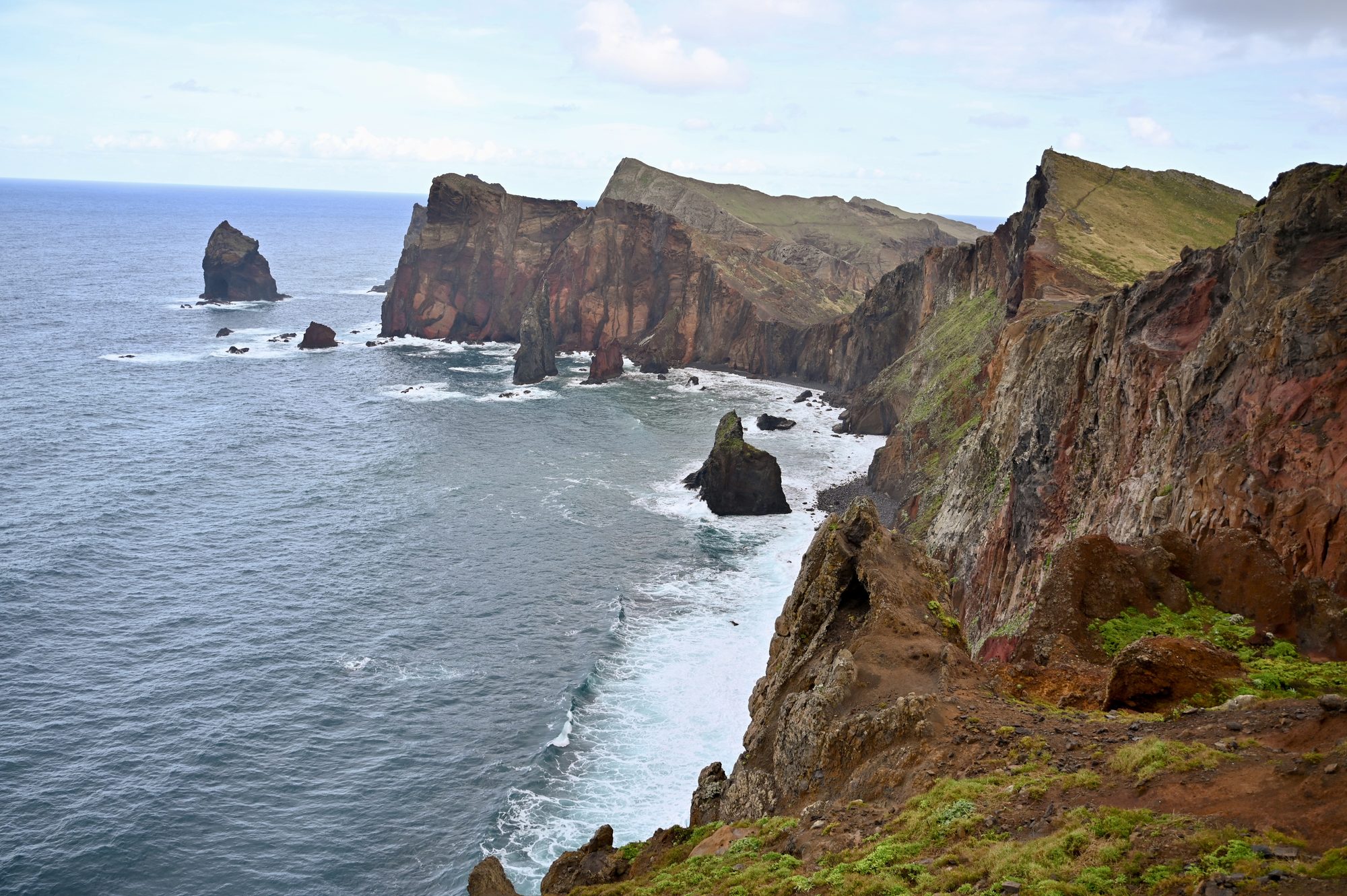 Red and brown sea cliffs and rock stacks rising from a grey sea.