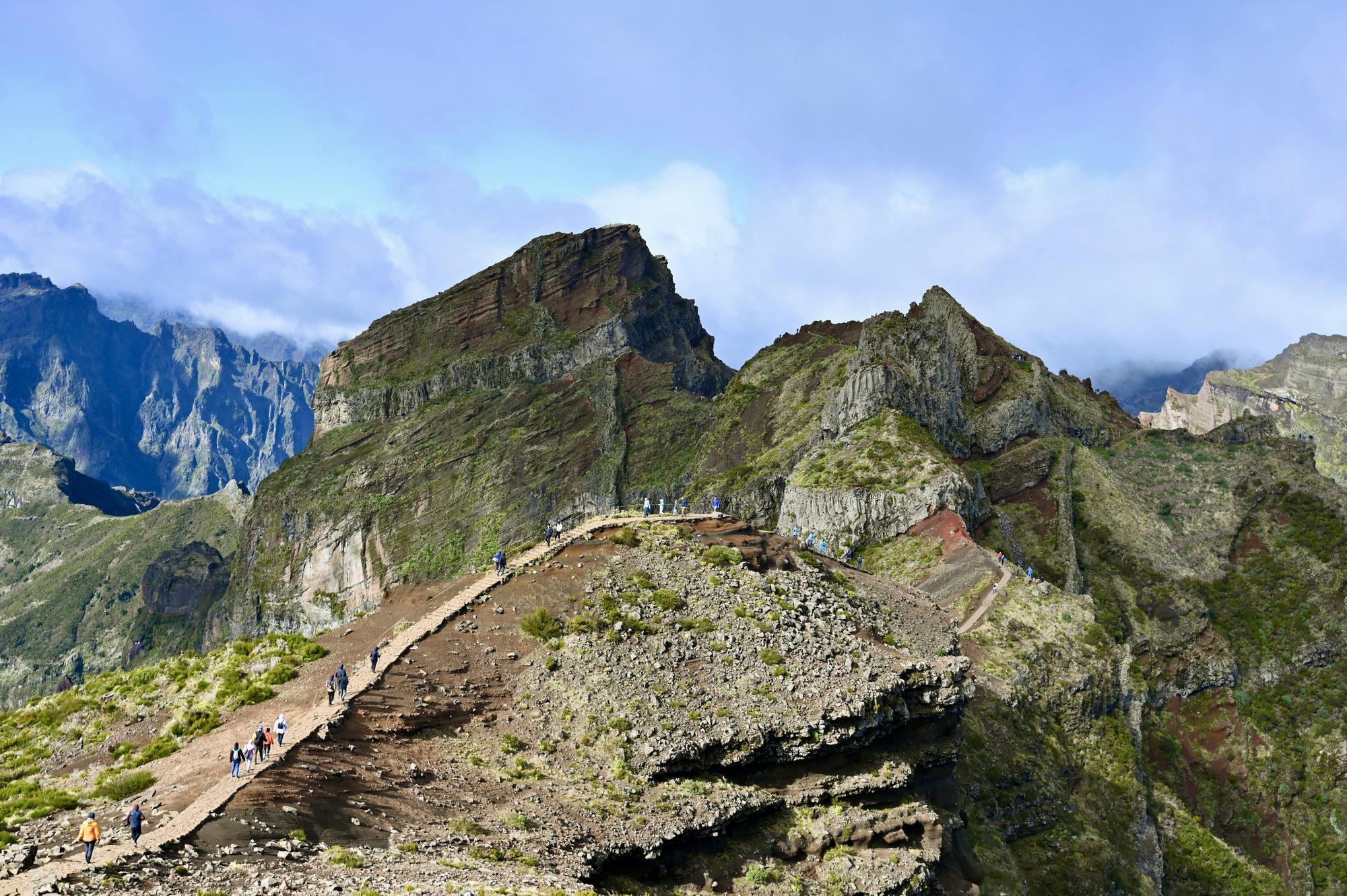 A paved path running along a narrow ridge towards a craggy peak, with tiny hikers spread out along it.