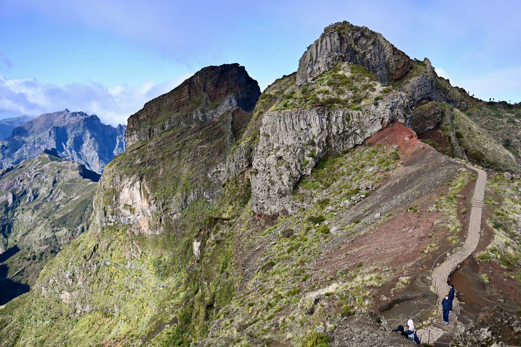 The trail climbing a red-earth ridge towards rocky green summits under a blue sky.