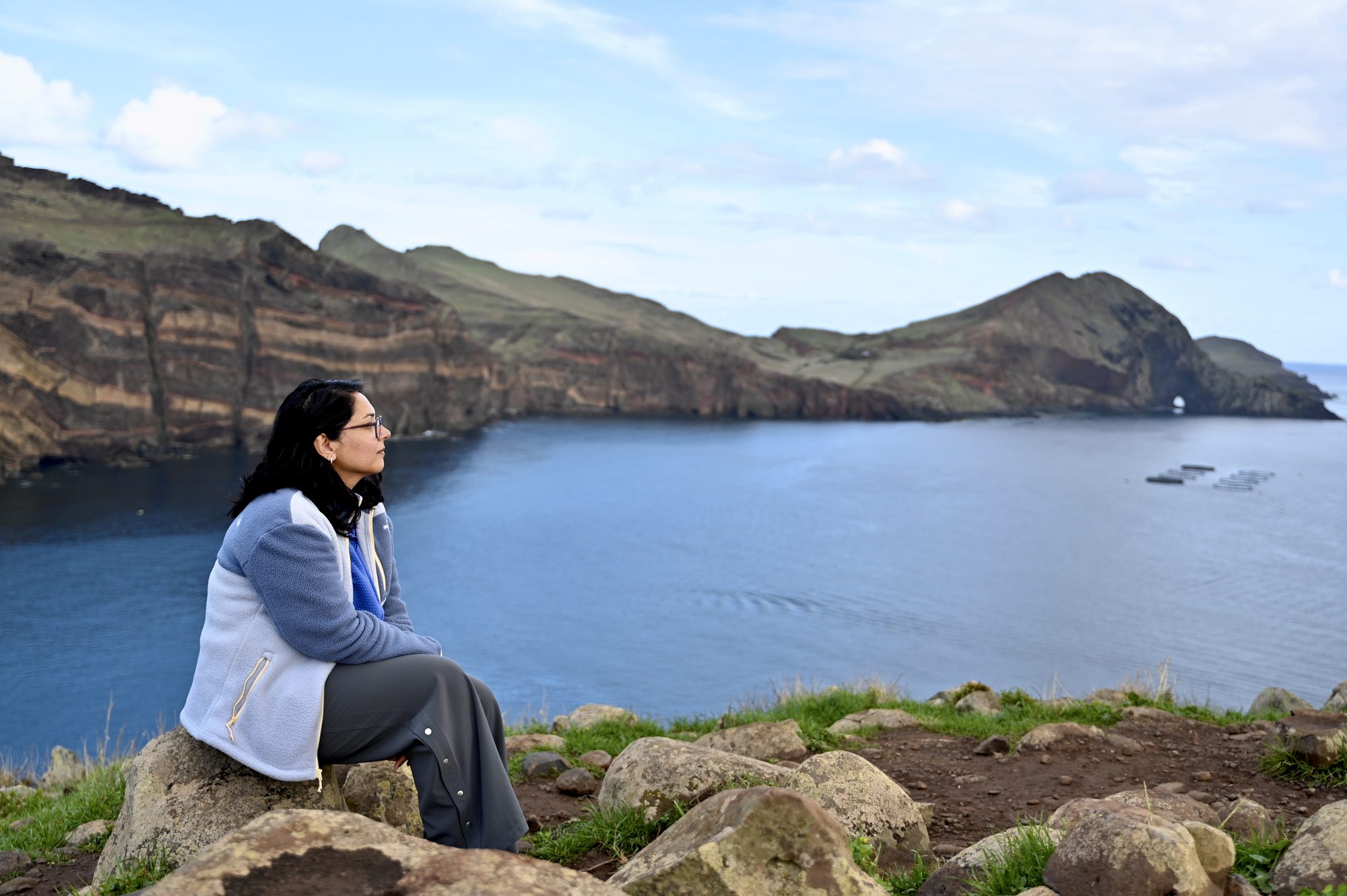 One of us sitting on a rock, looking out over a calm blue bay to a headland with a natural arch.