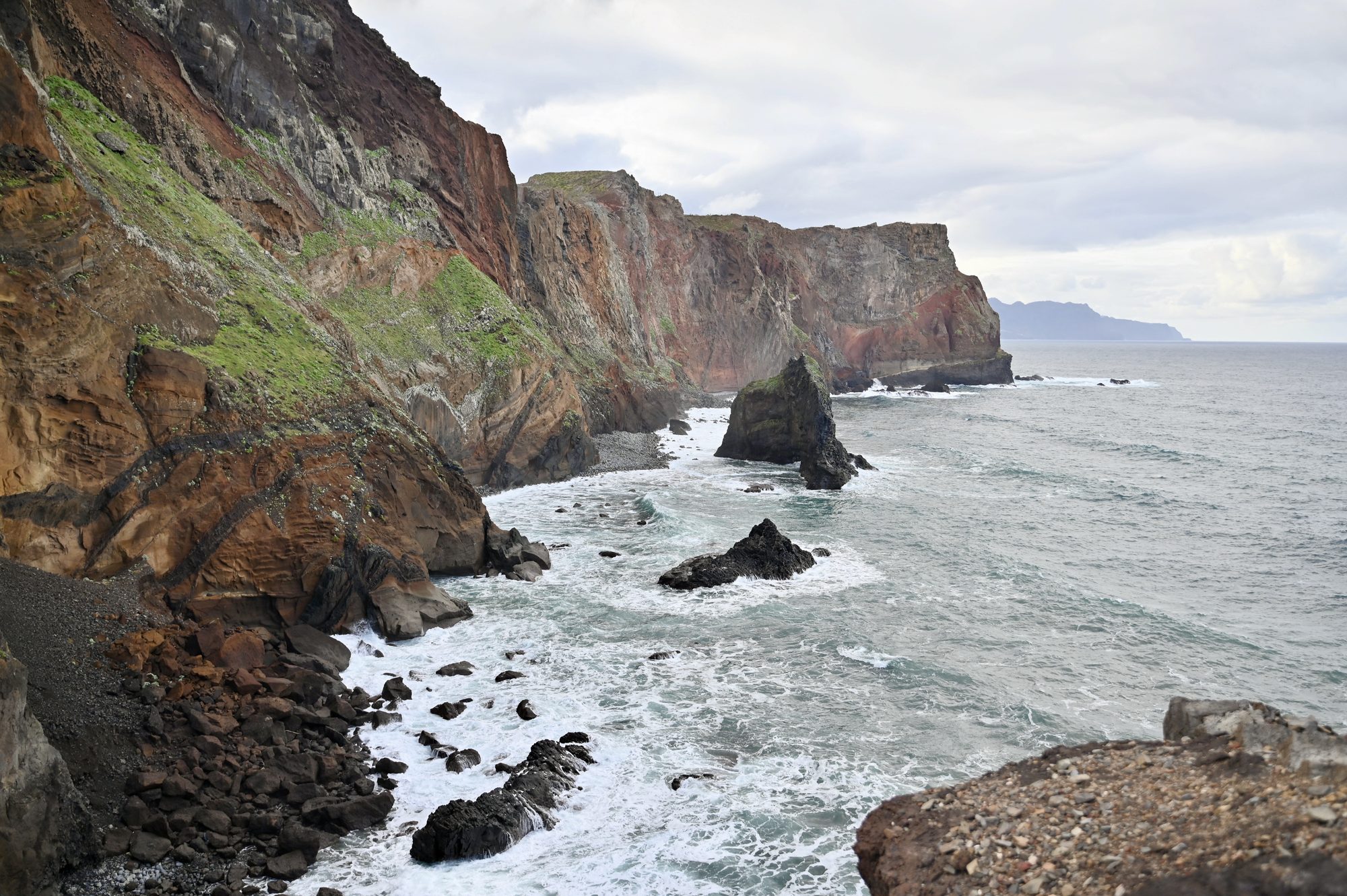Waves breaking against dark rocks below tall red and green cliffs.