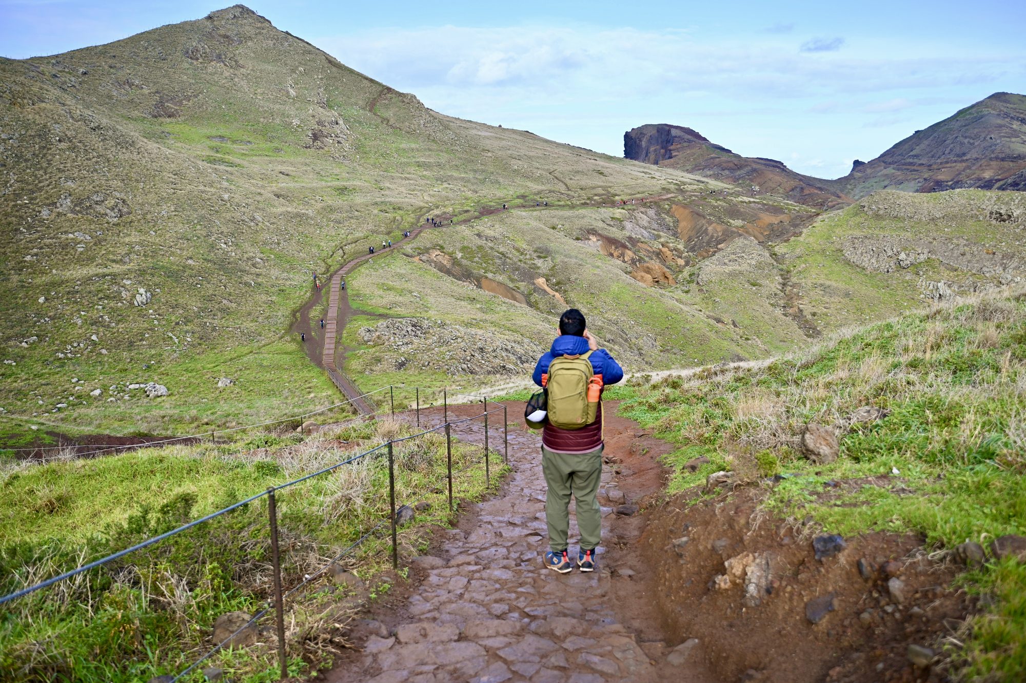 His dad walking away down a stone path with a railing, across green hills towards red cliffs.