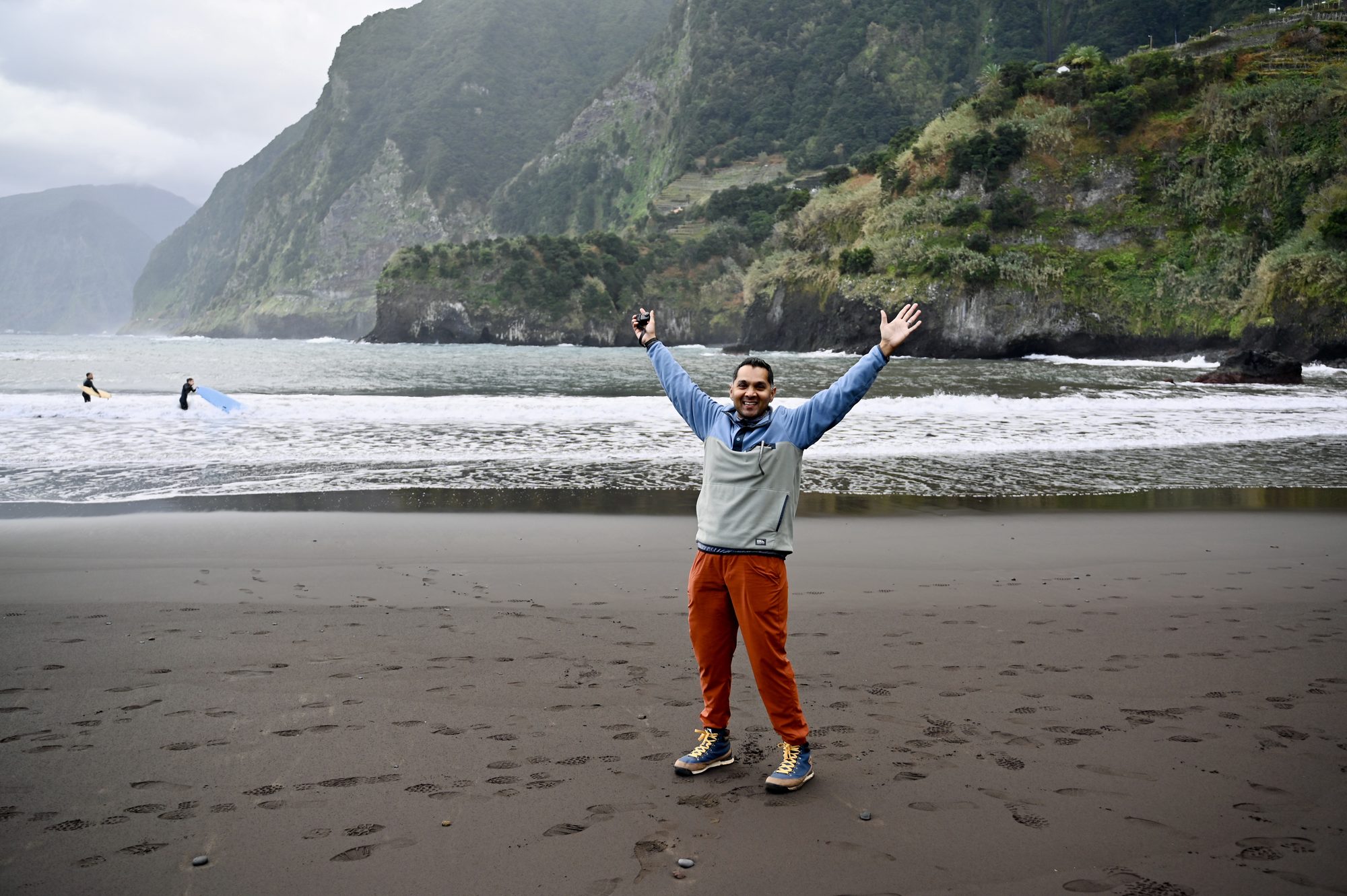 His dad with arms raised on the wide black-sand beach, with surfers in the waves and green cliffs behind.
