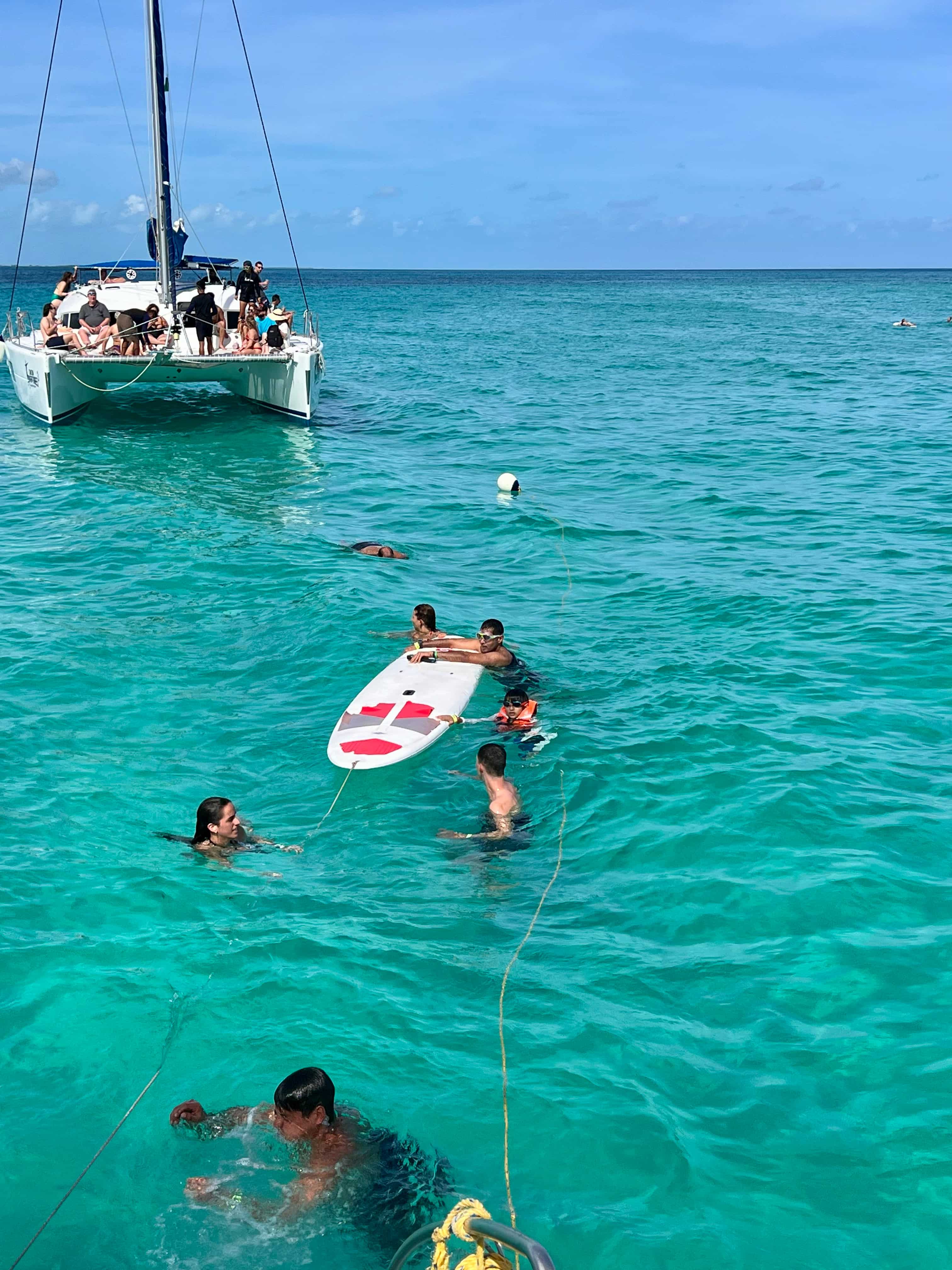 People in clear turquoise waters around a paddle board with a catamaran in the background