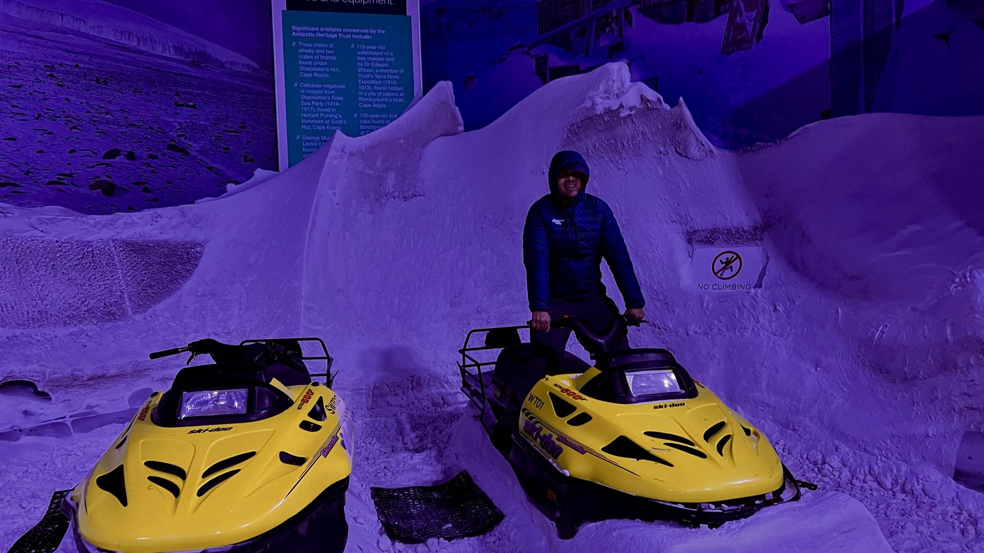 His dad in a padded jacket and hood beside two yellow snowmobiles in a snowy room lit blue.