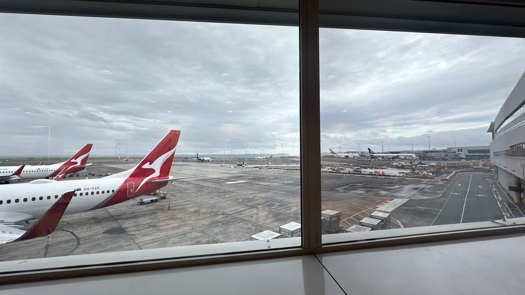 Two Qantas planes parked on a grey, overcast apron, seen through the terminal windows at Auckland airport.