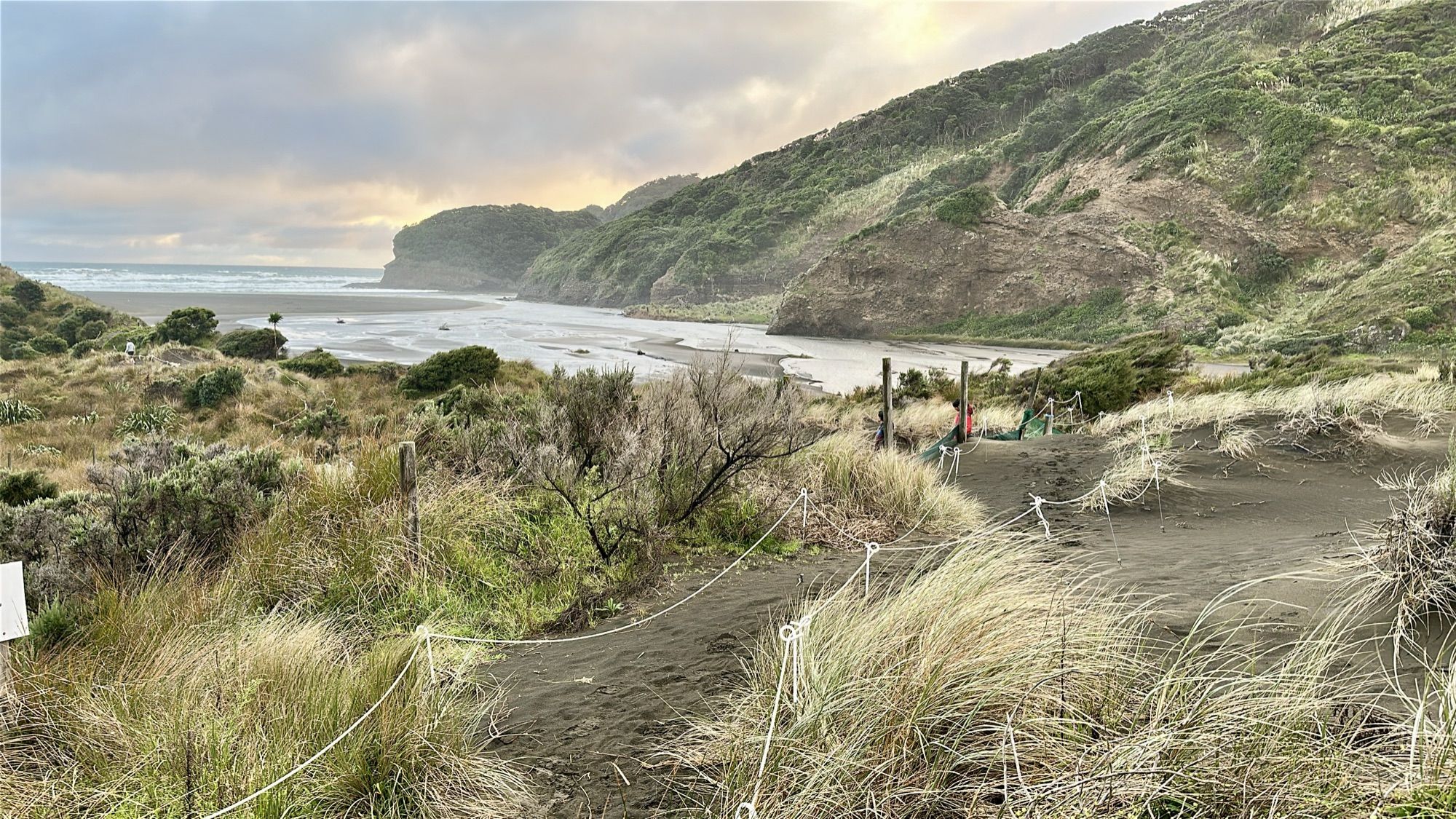 A sandy track through grassy dunes towards a wide beach and steep green hills, in late-afternoon light.