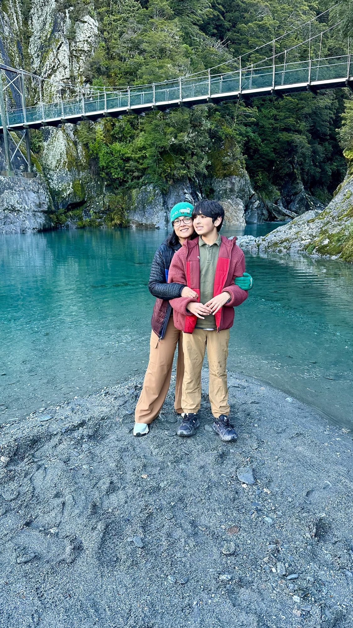 Our son and I hugging on a gravel bank beside the turquoise Blue Pools, with a swing bridge above.