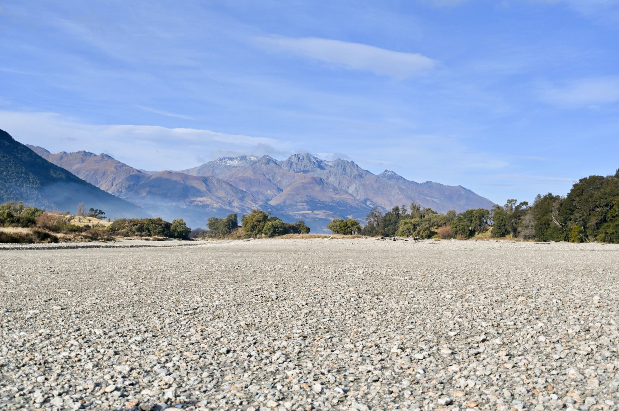 A wide, pale stony riverbed stretching towards forested hills and snowy peaks.