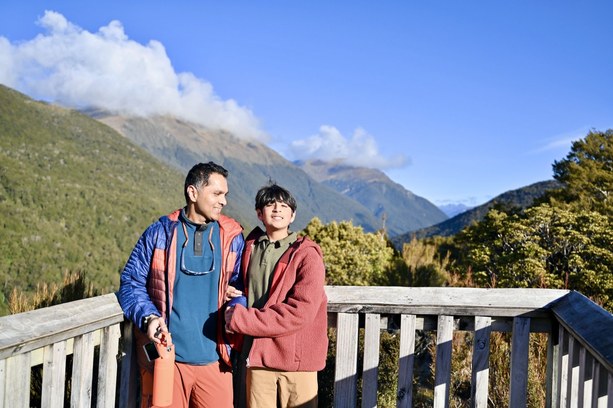 Our son and his dad at the wooden railing of the lookout, with forested mountains and clouds behind them.