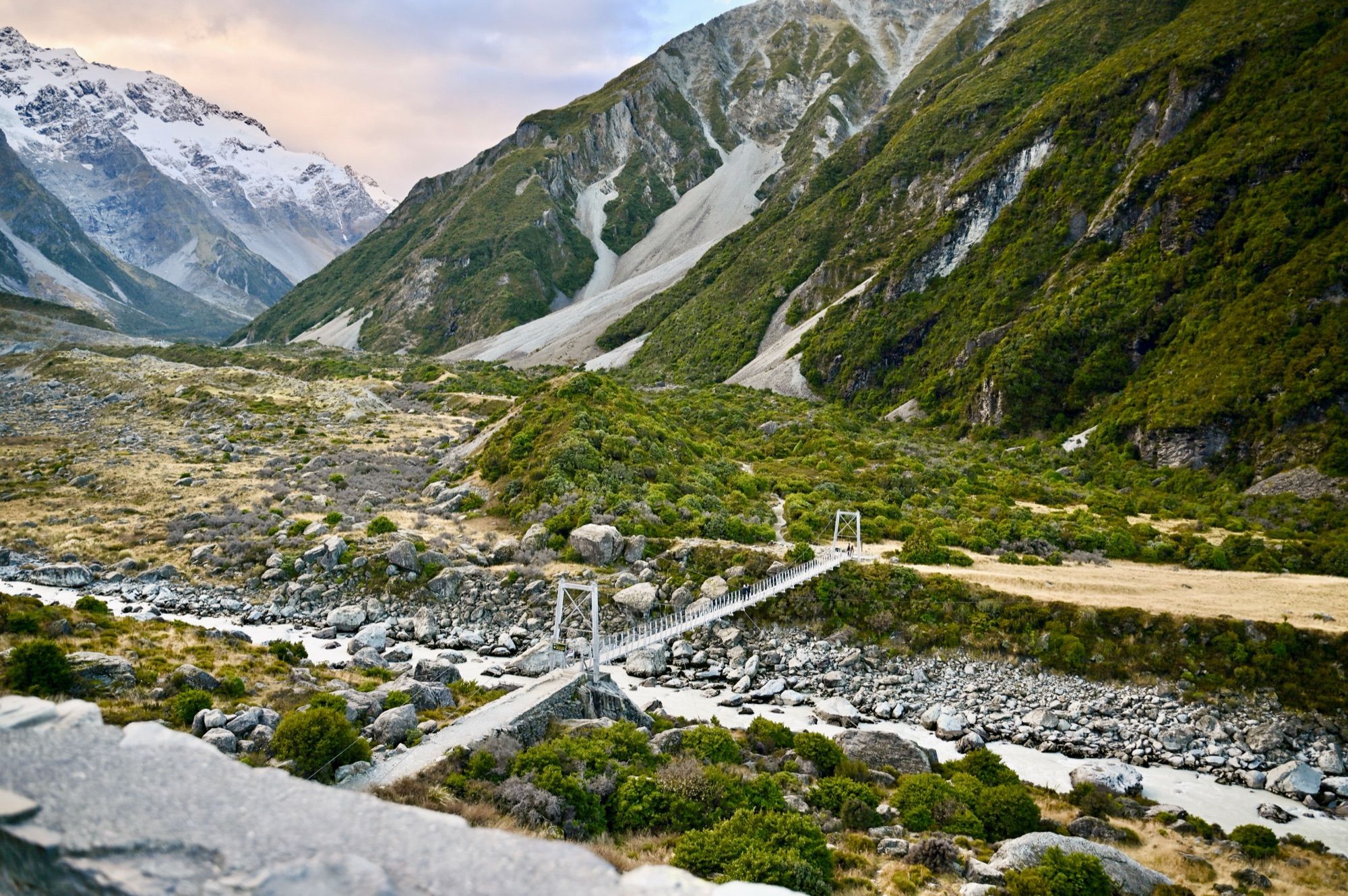 A swing bridge crossing a rocky glacial river below steep green and grey mountainsides.