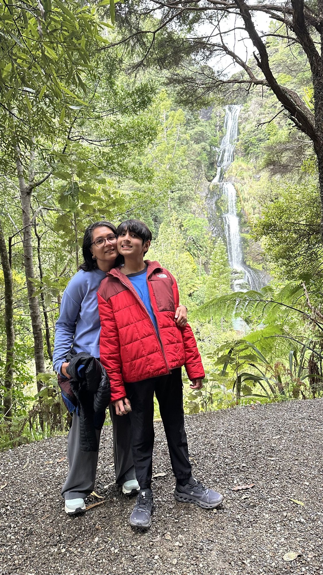 Our son and I standing together on the trail, with the long white ribbon of Kitekite Falls in the rainforest behind us.