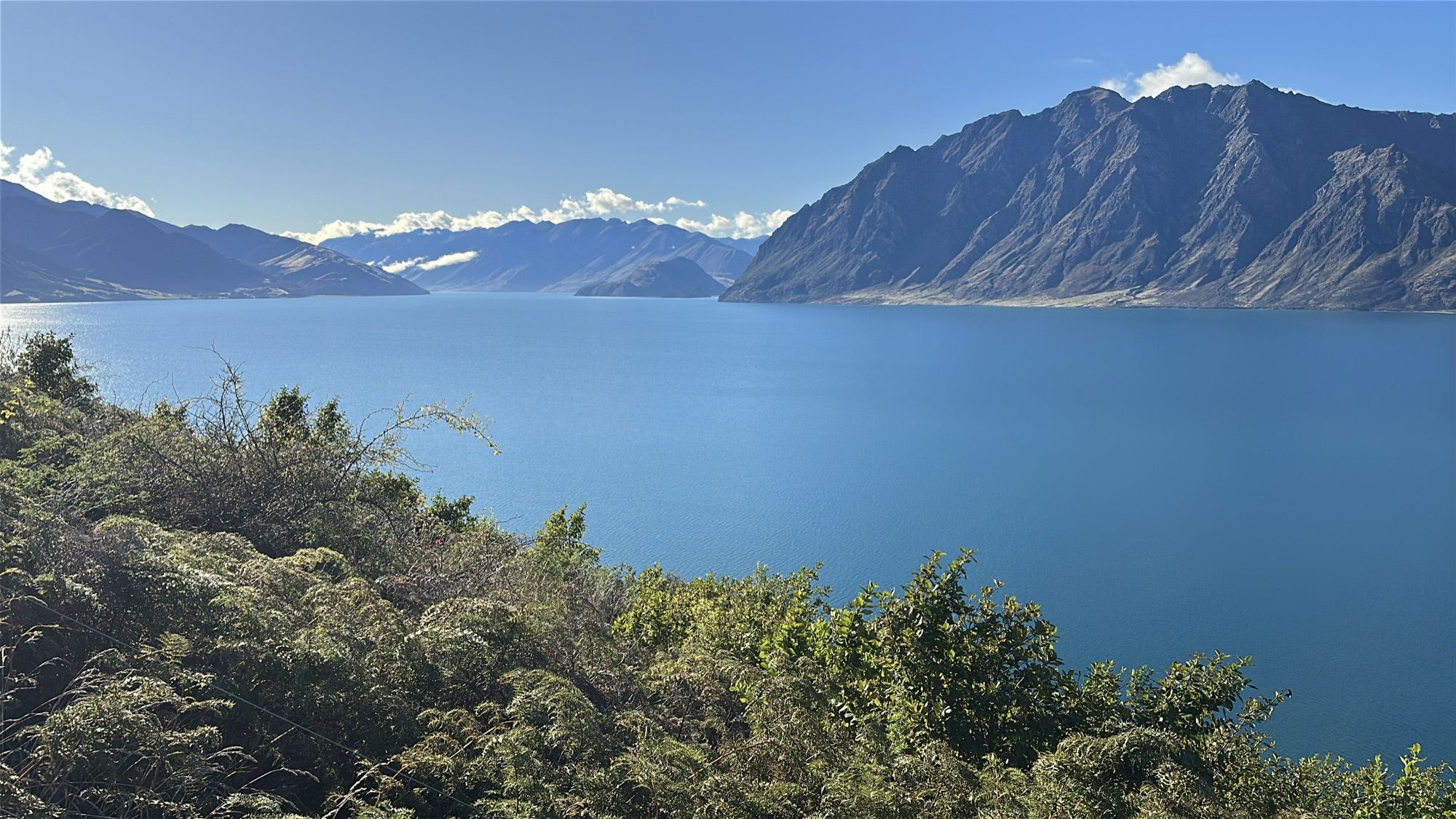 A deep blue lake framed by bush in the foreground and steep brown mountains, with clouds on distant peaks.