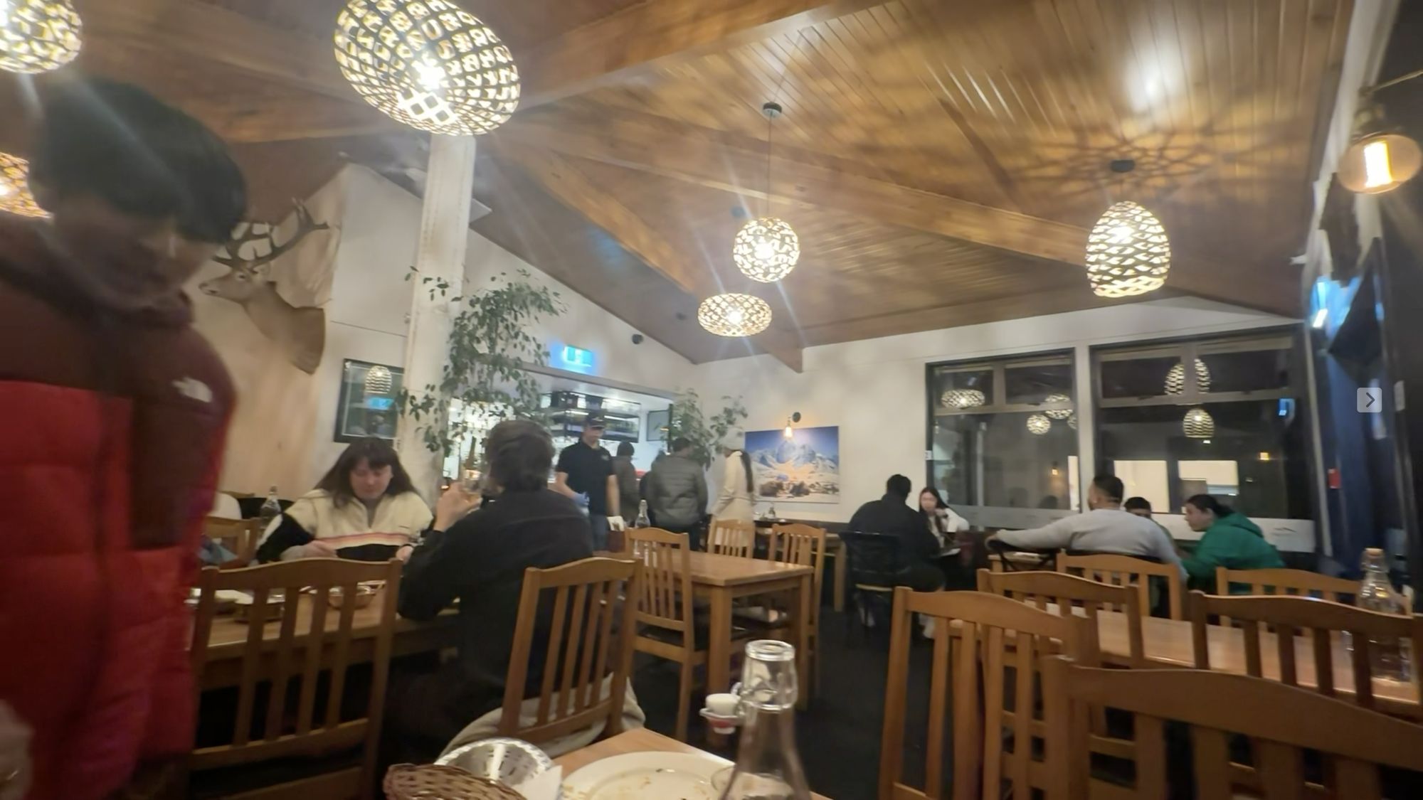 A cosy dining room with a wood-panelled ceiling, glowing pendant lights and diners at wooden tables.