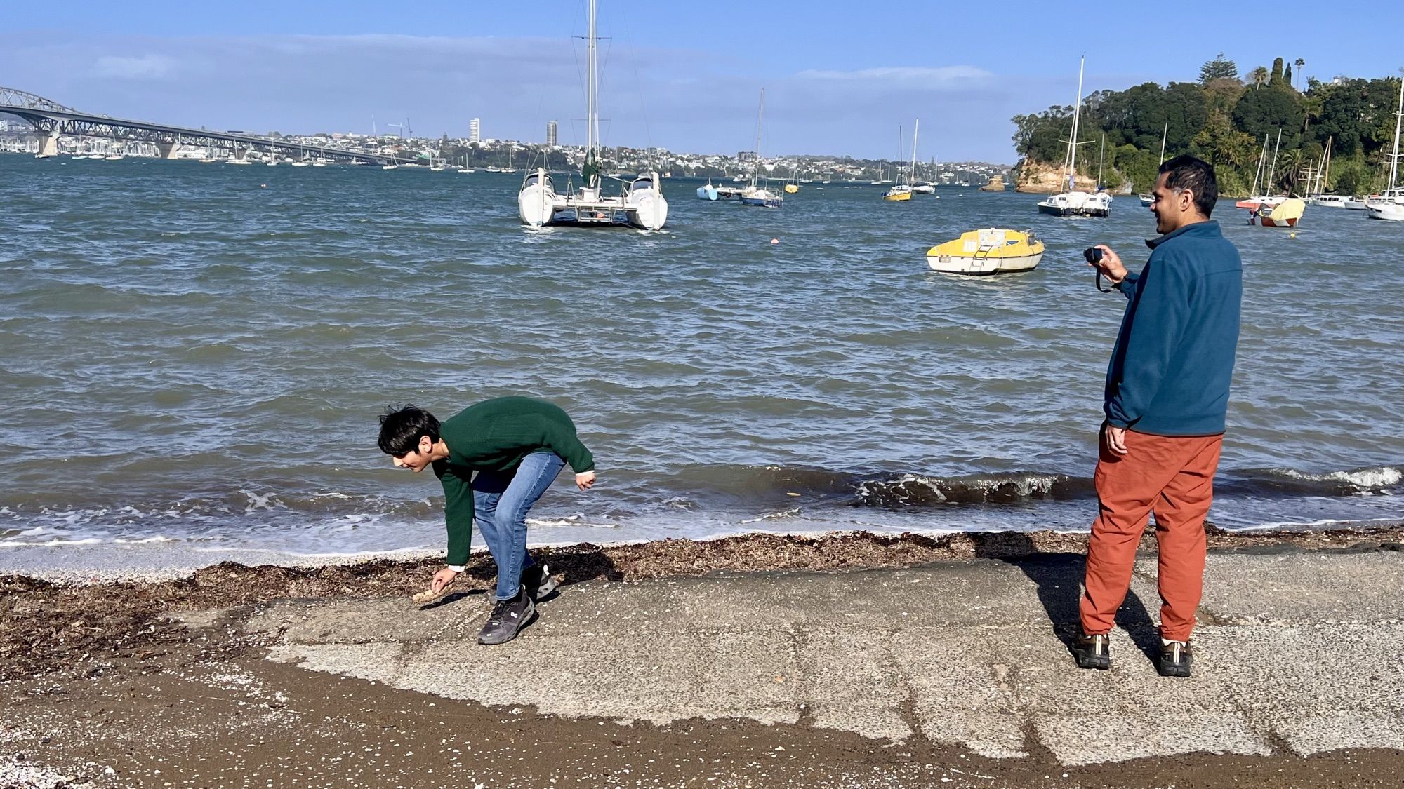 Our son bending down at the water’s edge while his dad takes a photo, with moored boats and the Harbour Bridge behind them.