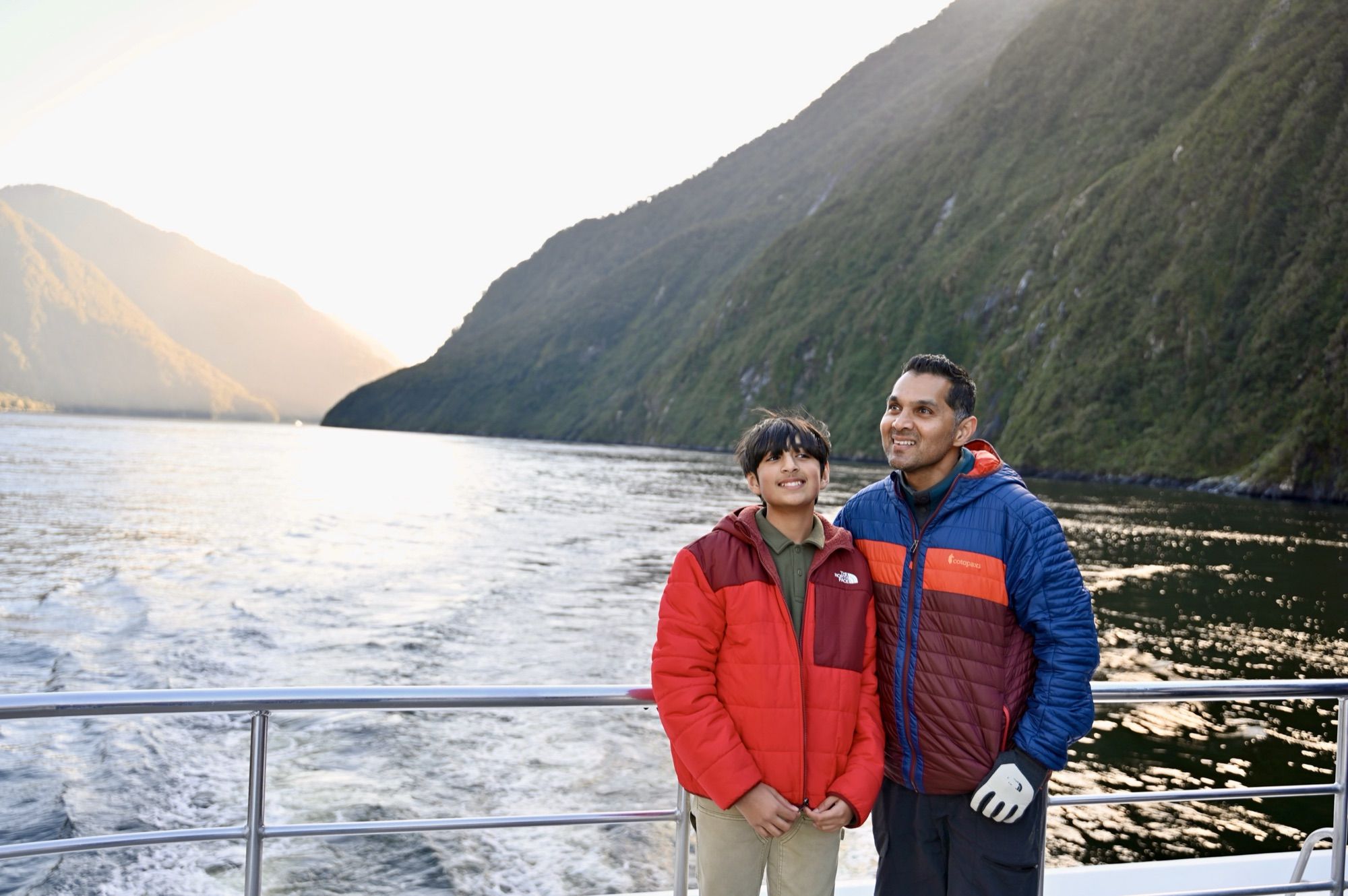 Our son and his dad smiling at the stern of the boat, with the fiord and the boat’s wake behind them.