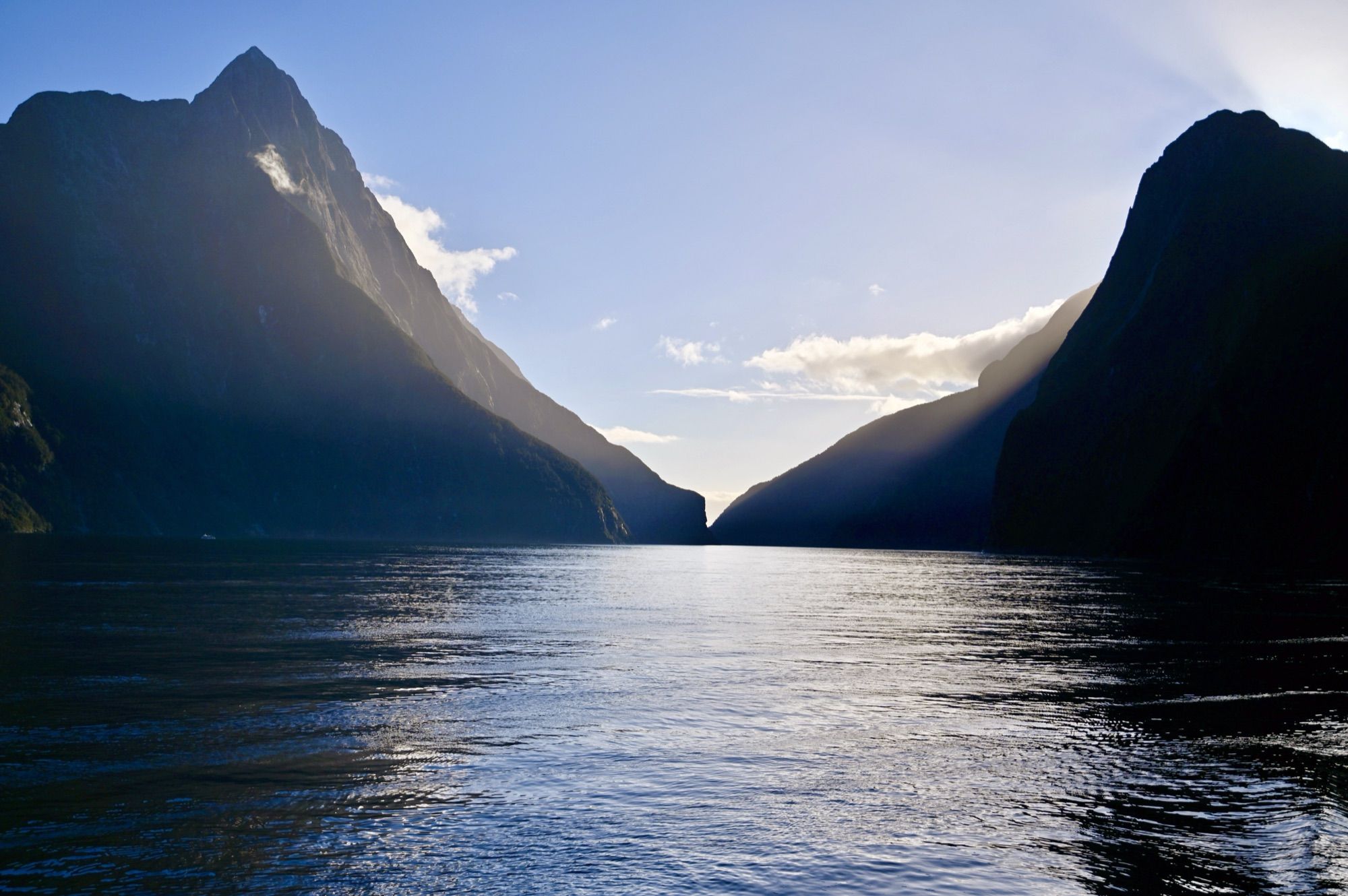 Mitre Peak and the neighbouring mountains in silhouette against a bright sky, above the shimmering water of Milford Sound.