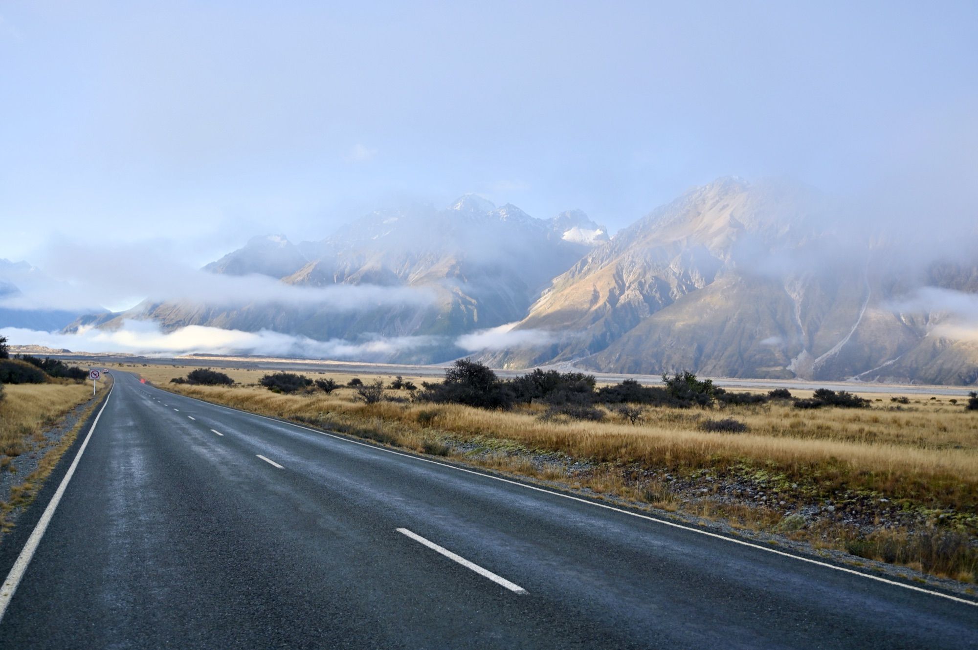 Low cloud lifting off snowy mountains beyond a wet, empty road across a tussock plain.