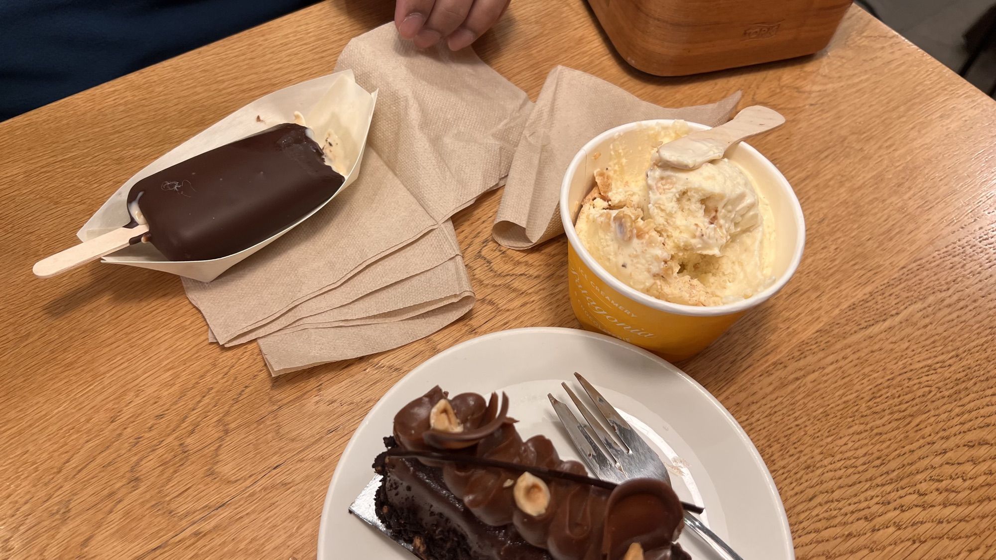 A chocolate-dipped ice cream bar, a cup of ice cream and a slice of chocolate hazelnut cake on a wooden table.