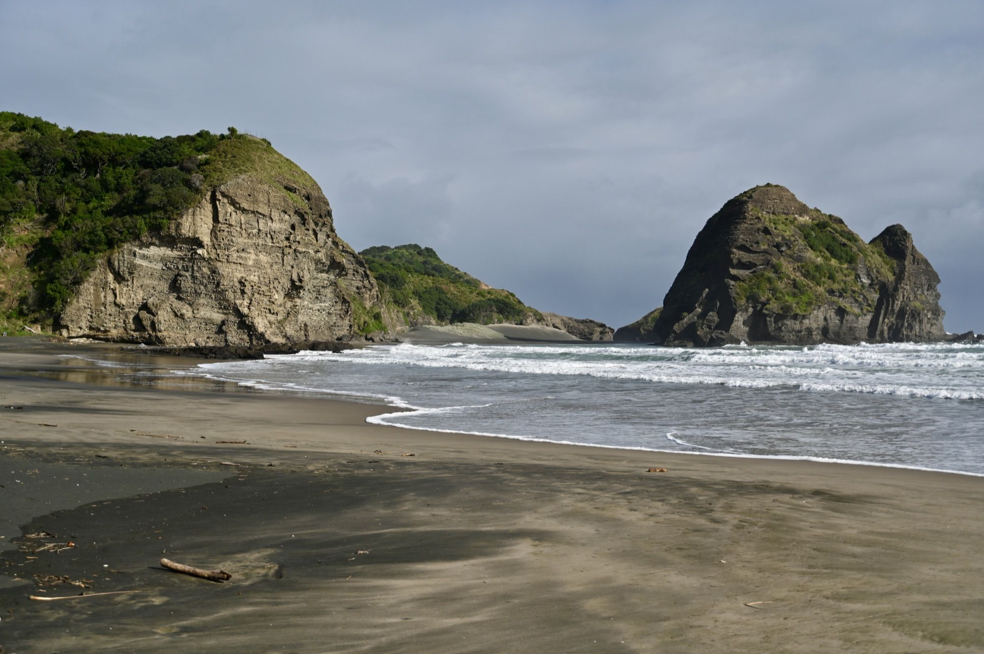 A black-sand beach with rolling surf, framed by a headland on the left and Lion Rock and smaller rocks on the right.