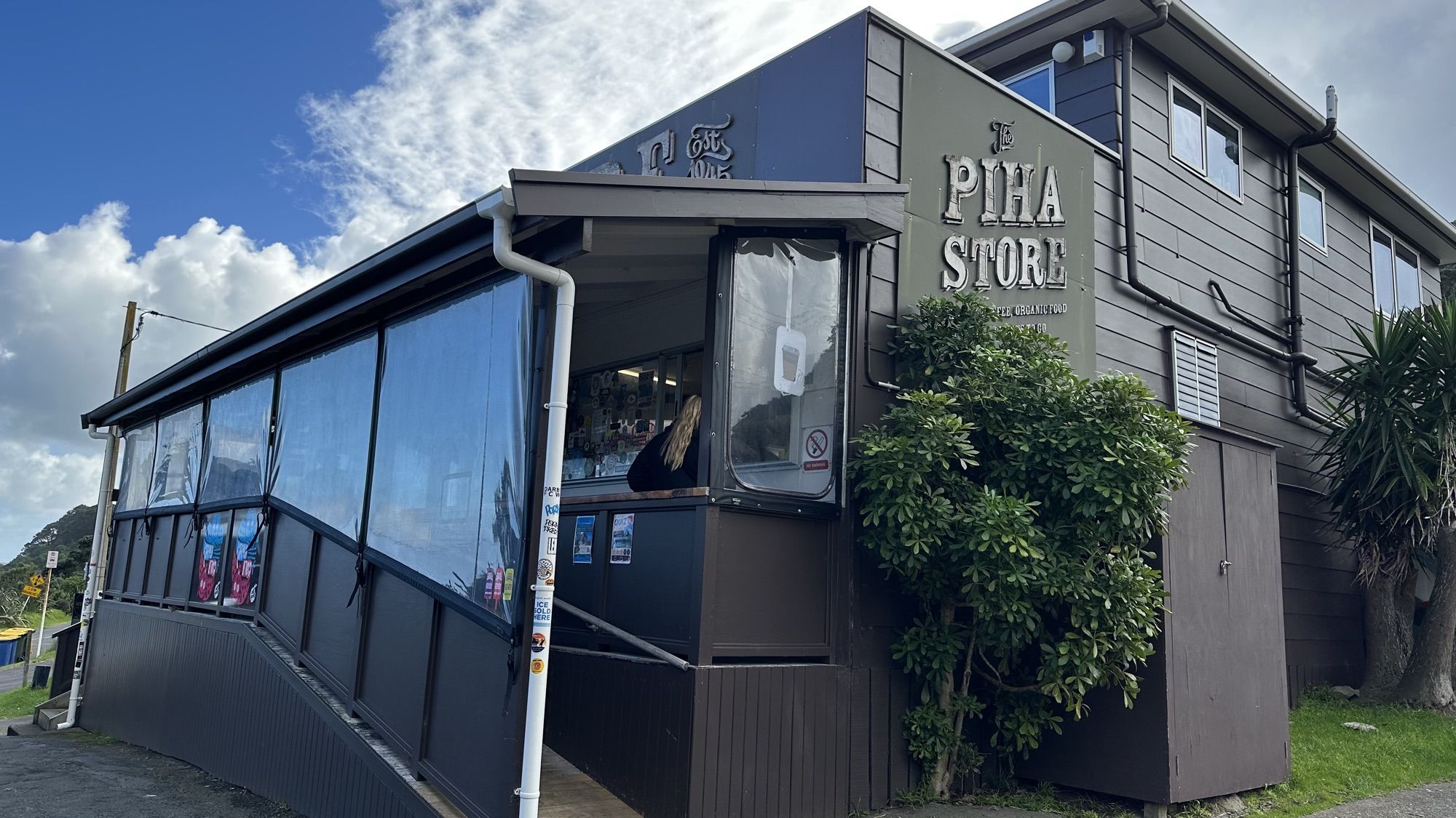 The dark wooden front of the Piha Store, with windows along a ramp and a bright blue sky above.