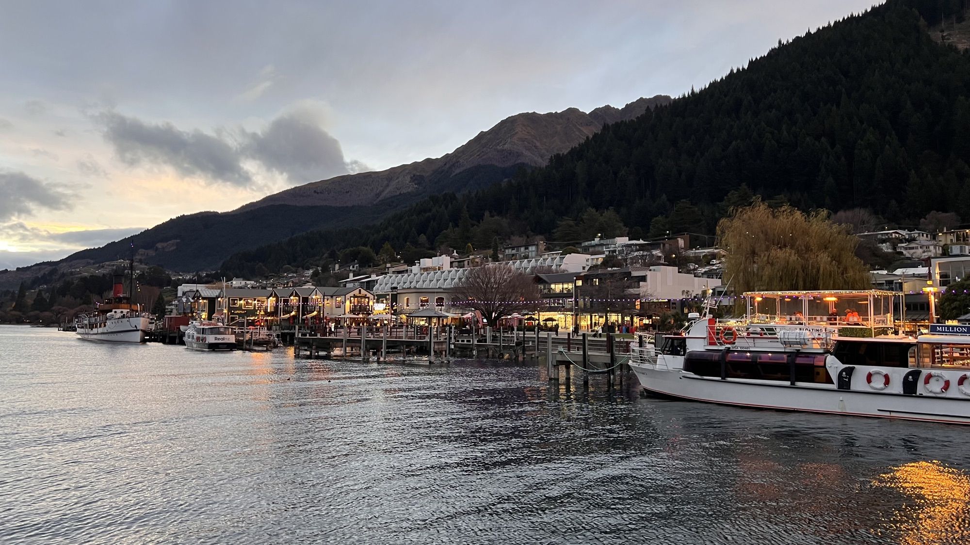Queenstown’s lakefront lit up at dusk, with boats moored at the wharf and dark mountains behind.
