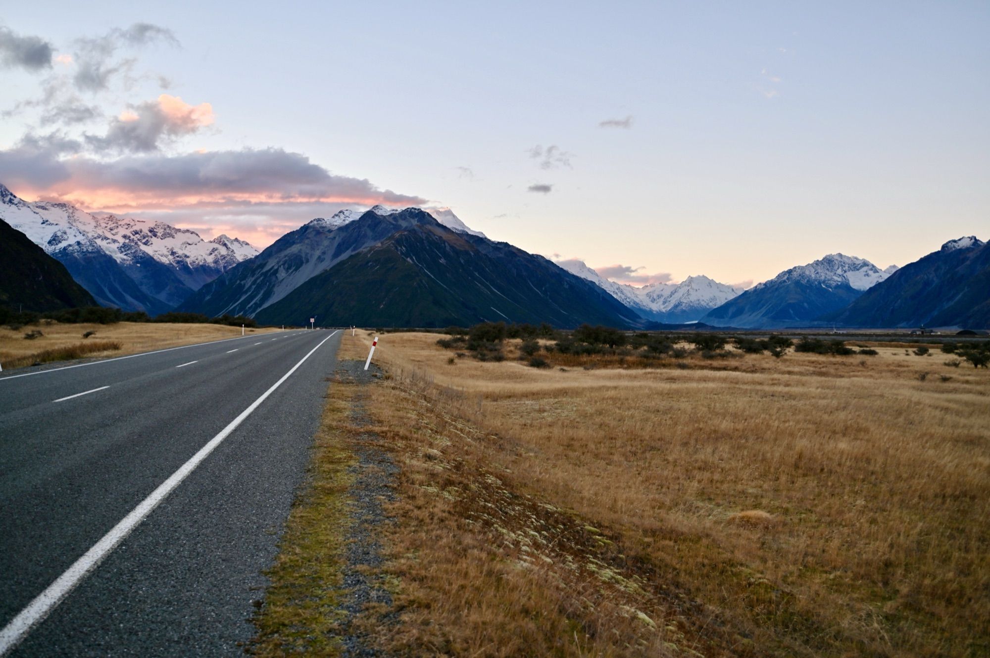 A straight road leading towards snow-capped mountains across a golden tussock plain, with pink clouds at sunset.