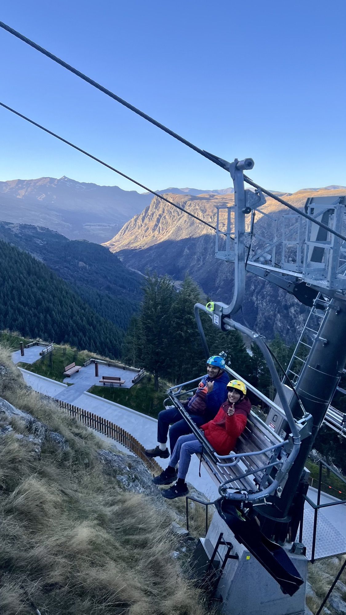 Two of us in helmets riding the chairlift back up to the luge track, with mountains and a valley behind.