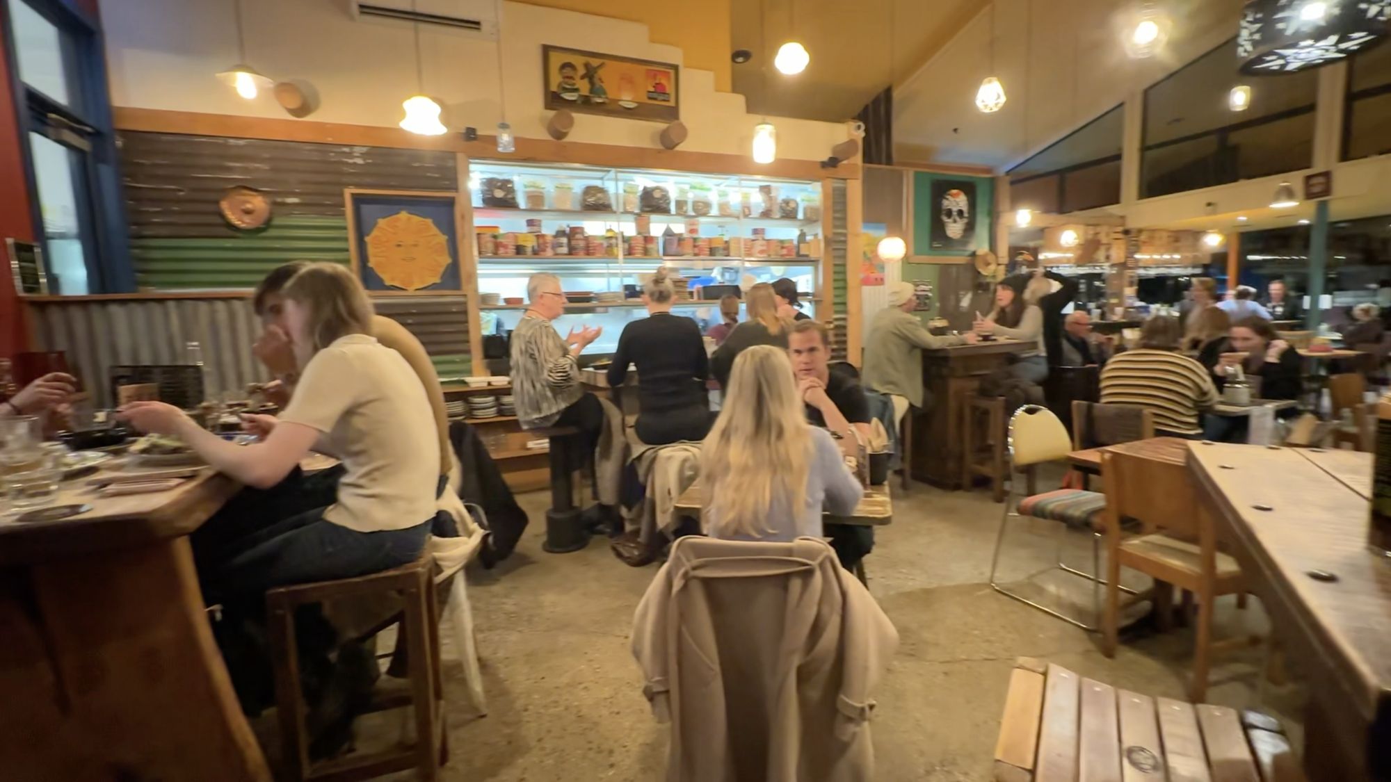 A busy, warmly lit restaurant with diners at wooden tables and a colourful shelf of jars behind the bar.