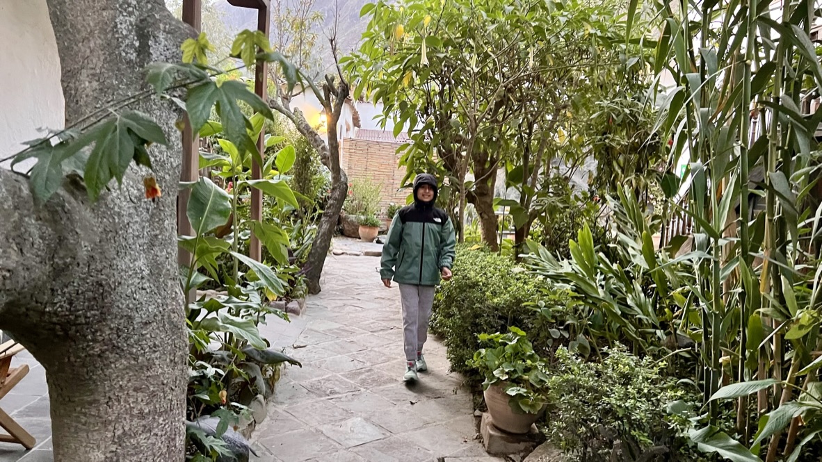 Boy in green and black jacket with hood smiling and walking towards the camera. The pathway is surrounded by potted plants and small trees. The pathway is paved with tile stones